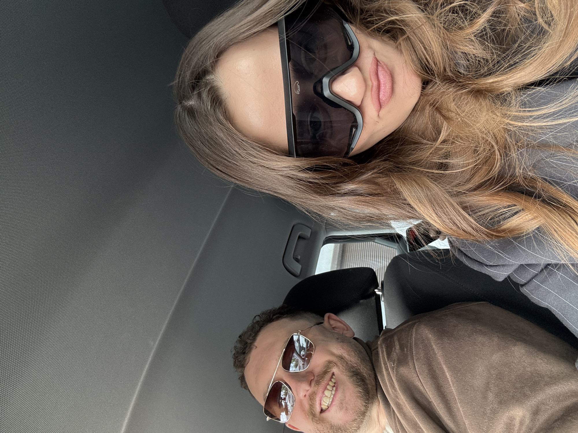 A selfie-style photograph captures a man and a woman inside a vehicle. The woman, positioned in the upper portion of the frame, has long, wavy brown hair and is wearing large, black sunglasses. Her lips are visible. The man, located in the lower part of the image, has short, curly brown hair and a beard. He is wearing sunglasses with brown lenses and is smiling, revealing his teeth. The interior of the vehicle is visible, including the dashboard and seat. Based on available information, the location is within a vehicle in Krasnoyarsk, Russia.