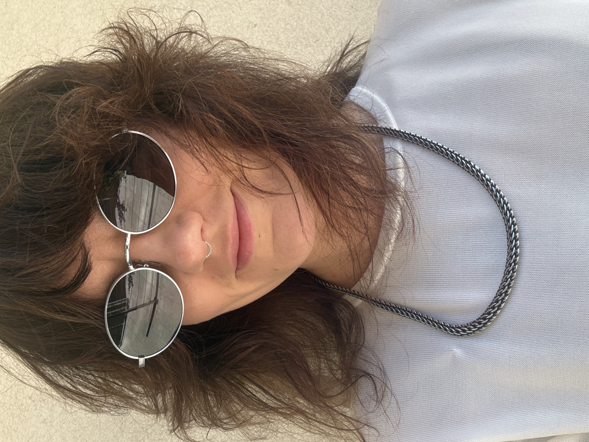 A person is seen from an overhead perspective, angled. The individual has brown hair, fair skin, and is wearing round sunglasses with a silver frame. A small nose ring is visible on the left nostril. The person is wearing a light-colored garment, and a thick silver chain necklace is draped around the neck. The background appears to be a light-colored surface. The setting appears to be outdoors, but location information is not discernible from the image. Therefore, identifying the location as the city of Budva, Montenegro is impossible.