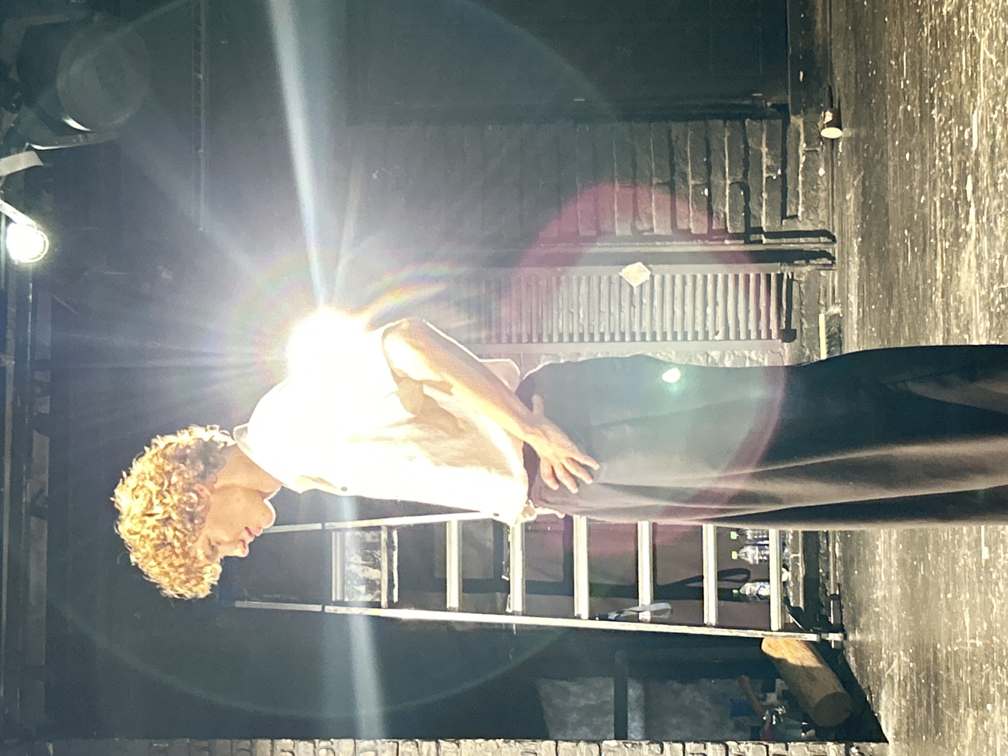 The image shows a person with curly blonde hair standing on a stage. The person is wearing a light-colored shirt and dark pants. They are leaning forward with their hands resting on their pants. The background is dark and appears to be a backstage area, with lighting equipment and a ladder visible. The scene is lit by a bright spotlight, which creates a strong glare and lens flare. There is no visible text in the image. The overall impression is one of a theatrical performance or rehearsal in progress, with a focus on the performer. The mood is somewhat dramatic due to the lighting.