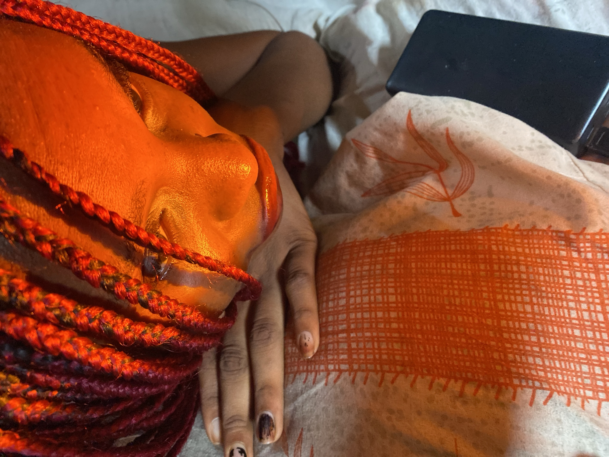 This media file is a close-up, overhead shot of a person lying down. The subject's face is illuminated by a warm, orange light, highlighting the contours of their cheek and nose. Red braids, interspersed with strands of darker color, frame the left side of their face and extend downwards. Their right hand rests on a white and orange patterned fabric, with some of the pattern resembling woven straw and delicate floral motifs. The person's fingernails have dark and light polish, with some smudging. To the right of the fabric, a dark, rectangular object, possibly a phone, is partially visible. The overall scene suggests a relaxed, intimate moment, potentially in a domestic setting. The lighting and intimate framing evoke a sense of calm and personal space. The location context provided is Iranla, Nigeria. There is no visible text in the image.