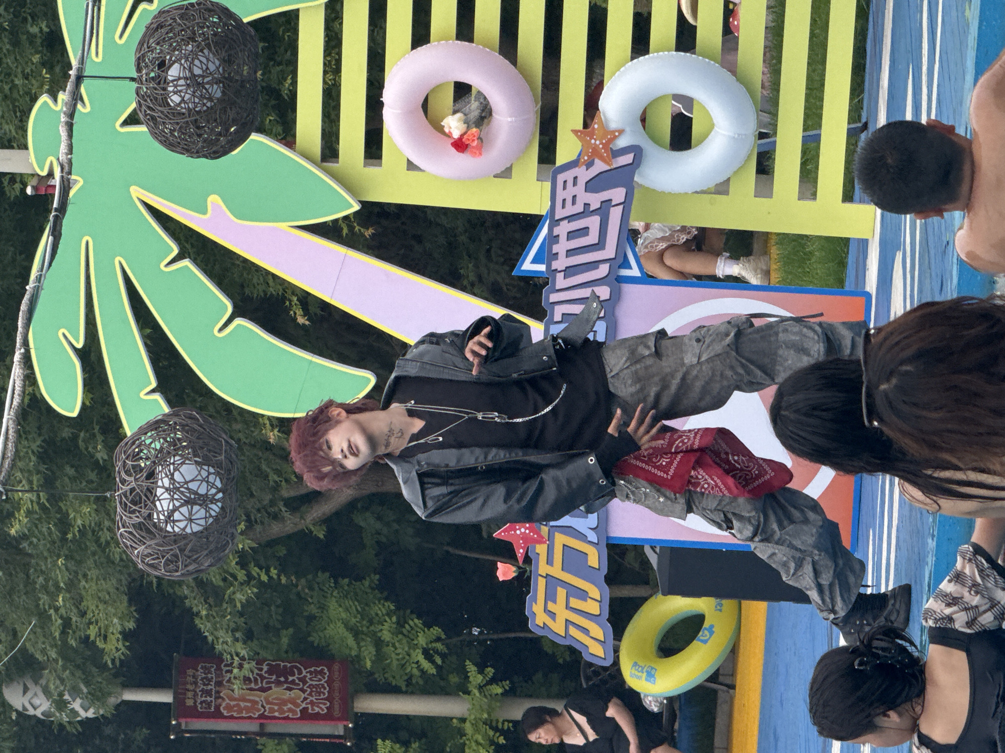 In the city of Taihe District, China, a young male with red-dyed hair and tattoos on his neck is posing in front of a colorful backdrop that resembles a tropical scene with a large palm leaf and a yellow, green, and pink striped structure. He is wearing a black leather jacket over a black v-neck shirt, camouflage cargo pants, and black boots. A red bandana is tucked into his pants. He is adorned with silver necklaces and a safety pin earring.
In the background, a woman in a black dress is seated on a bench, and other people are partially visible, some in what appears to be a pool. Two decorative woven spheres hang from above. There are inflatable rings, one yellow and one light blue, as well as a pink inflatable ring with small floral decorations. A sign with Chinese characters is also visible on the left side of the image. The overall scene suggests an outdoor recreational event or performance.