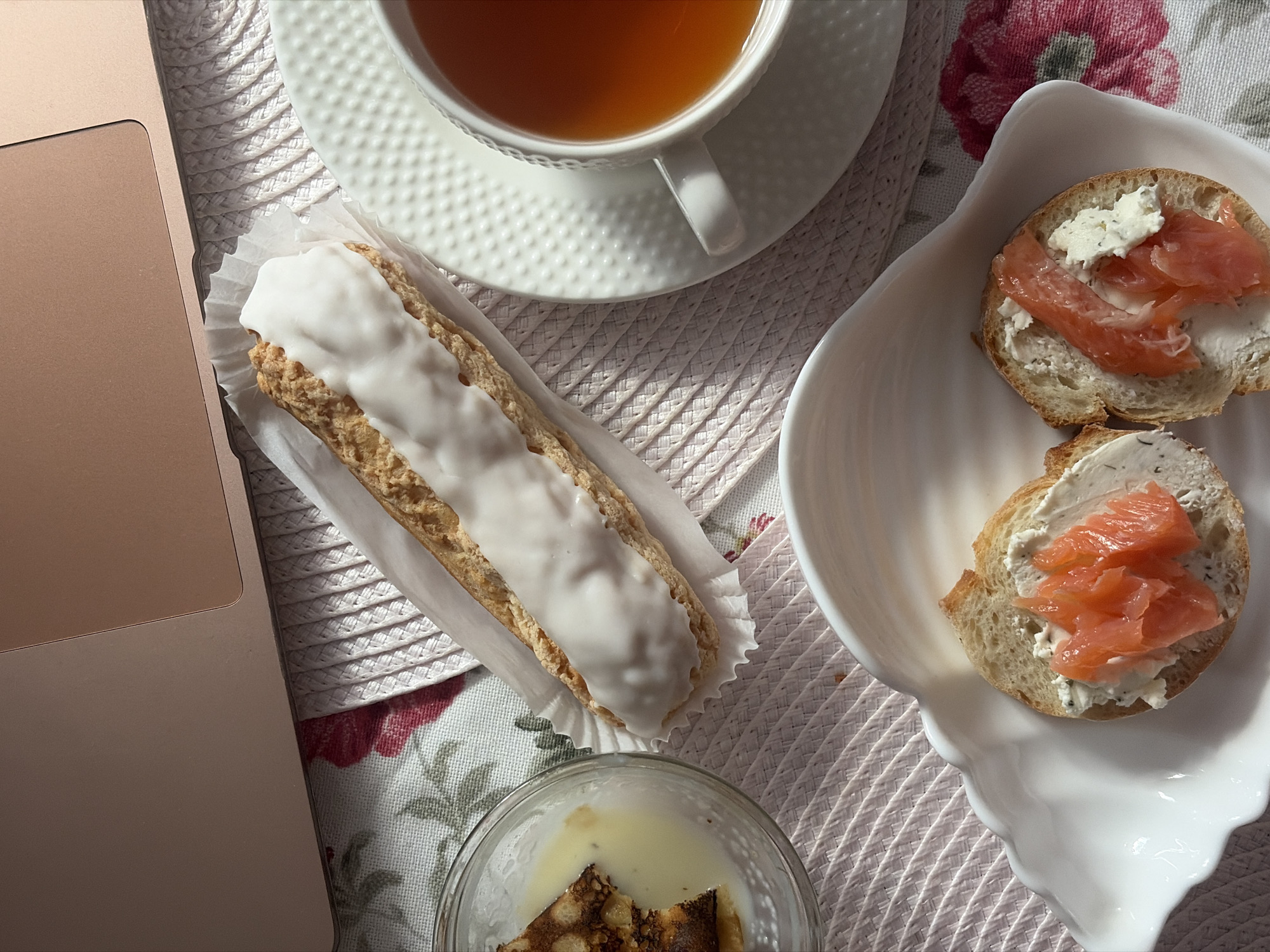 The image shows a still life on a surface with a floral pattern. A laptop is visible on the left side of the frame. A white teacup with tea is on a saucer, an eclair with white icing in a paper liner, and a small glass bowl with liquid and a dark, possibly burnt, substance at the bottom. A white decorative plate holds two pieces of bread topped with cream cheese and salmon. The image is taken from a high-angle perspective. There are no people visible. The presence of tea, pastries, and a laptop suggests a relaxed, possibly work-from-home setting. Given the items shown, it is not possible to determine if the location is Saint Petersburg, Russia.