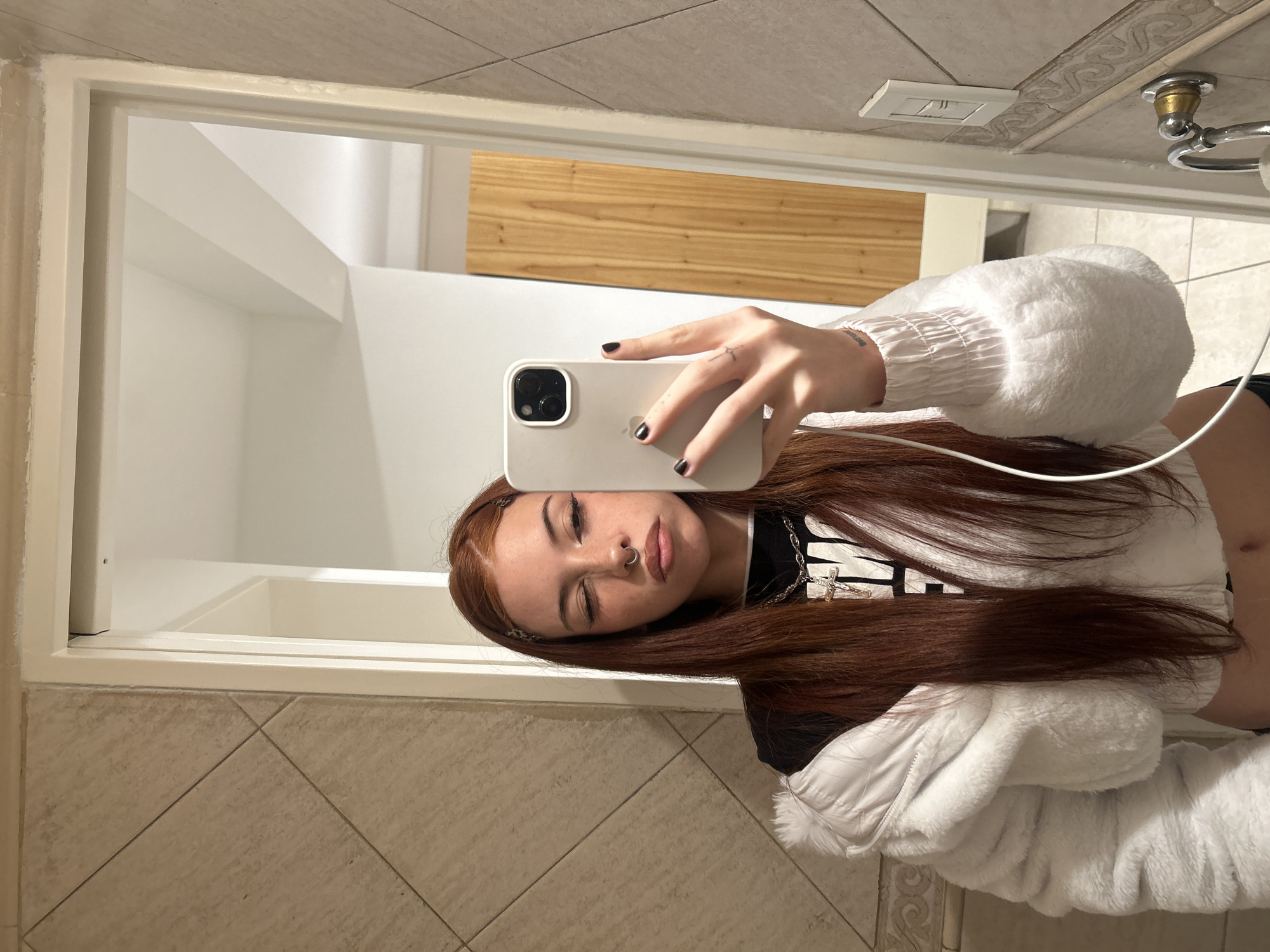 A young woman takes a selfie in a bathroom. She holds a white iPhone in her right hand, revealing her face. She has auburn hair, fair skin, and a pierced septum. Her hair falls across her shoulders and she wears a black top beneath a white, furry jacket. Her black nail polish is visible. The bathroom features tiled walls and a mirror reflecting the bathroom interior, including a bathtub, wooden panel, and light switch. The location in the city of Buenos Aires, Argentina cannot be determined from the image.