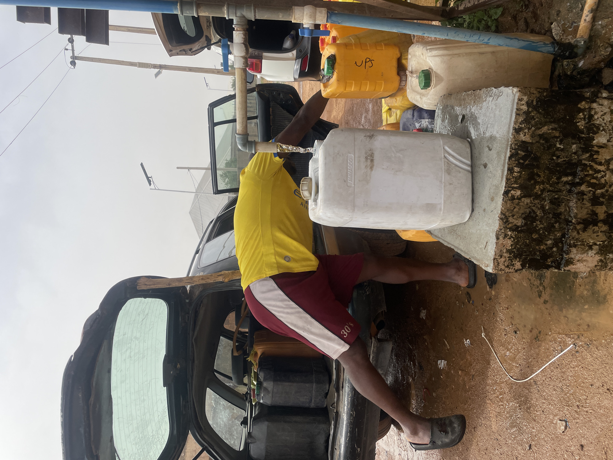 A person in a yellow shirt and burgundy shorts is filling a white container with water from a tap. Several yellow and white containers surround the person. The person is standing in a muddy area, and the back of a vehicle is visible, filled with black containers. The image shows the location is in Jos, Nigeria.