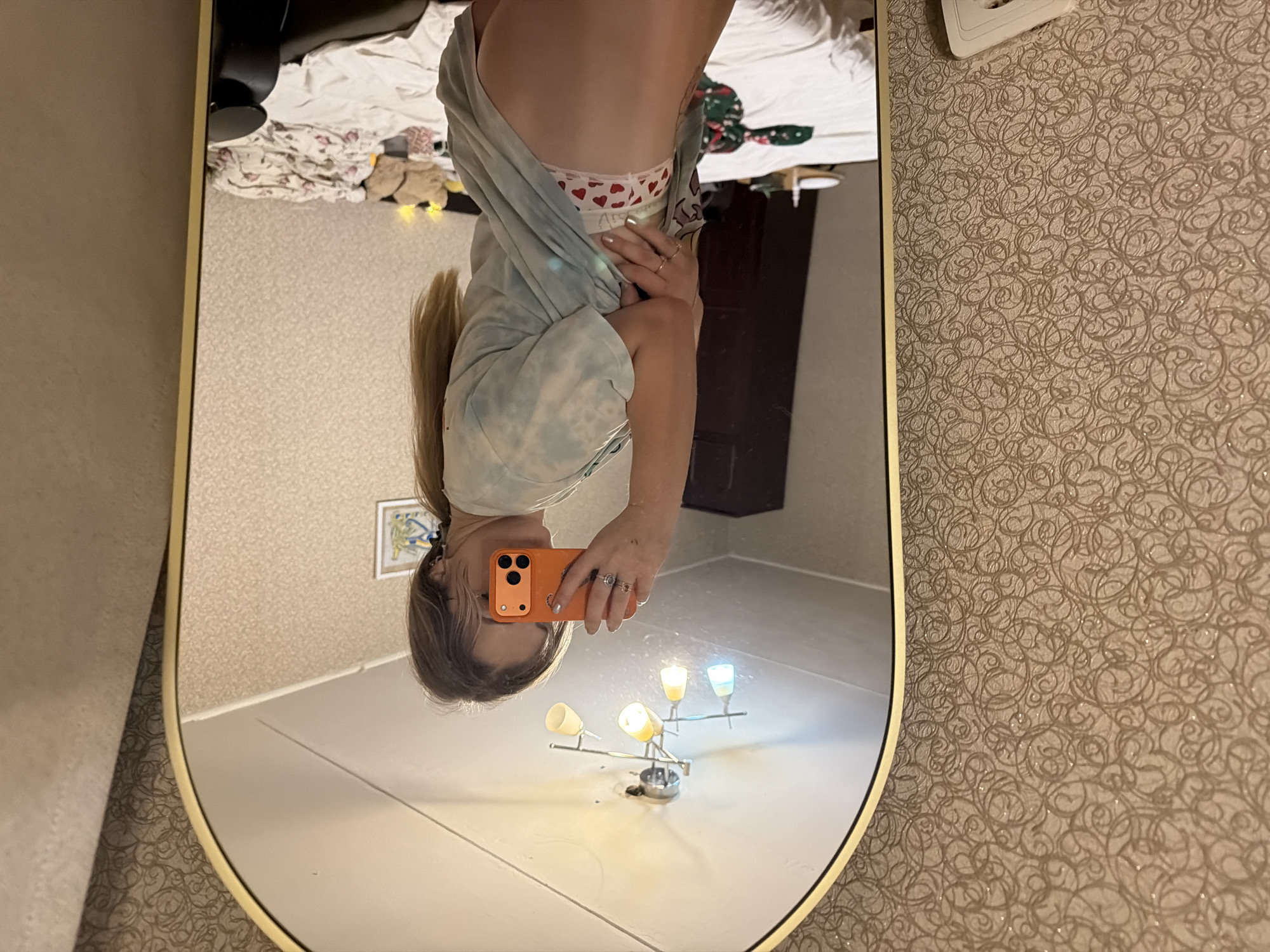 An upside-down mirror selfie captures a person in an indoor setting. The subject, likely female, has long, light-colored hair tied back, and is wearing a light-colored, possibly tie-dyed t-shirt and white underwear or shorts patterned with red hearts. The waistband of the underwear displays text that appears to read "ALS". Rings are visible on their fingers as they hold an orange smartphone with multiple camera lenses to take the picture.
The reflection in the oval-shaped mirror, framed in gold or brass, shows an unmade bed with white sheets and some clothing or fabric. A small, light-colored stuffed animal or toy is visible near the bed. A dark brown piece of furniture stands in the background of the reflection. On the floor of the reflected room, a small lamp with three illuminated bulbs casts bright light. A framed picture or artwork hangs on a wall in the distance within the mirror.
Outside the mirror, the wall is covered with textured wallpaper featuring intricate swirling patterns in a light brown or beige hue. A white light switch or electrical outlet is also present on this patterned wall. The scene suggests a personal room, possibly a bedroom, illuminated by artificial lighting.