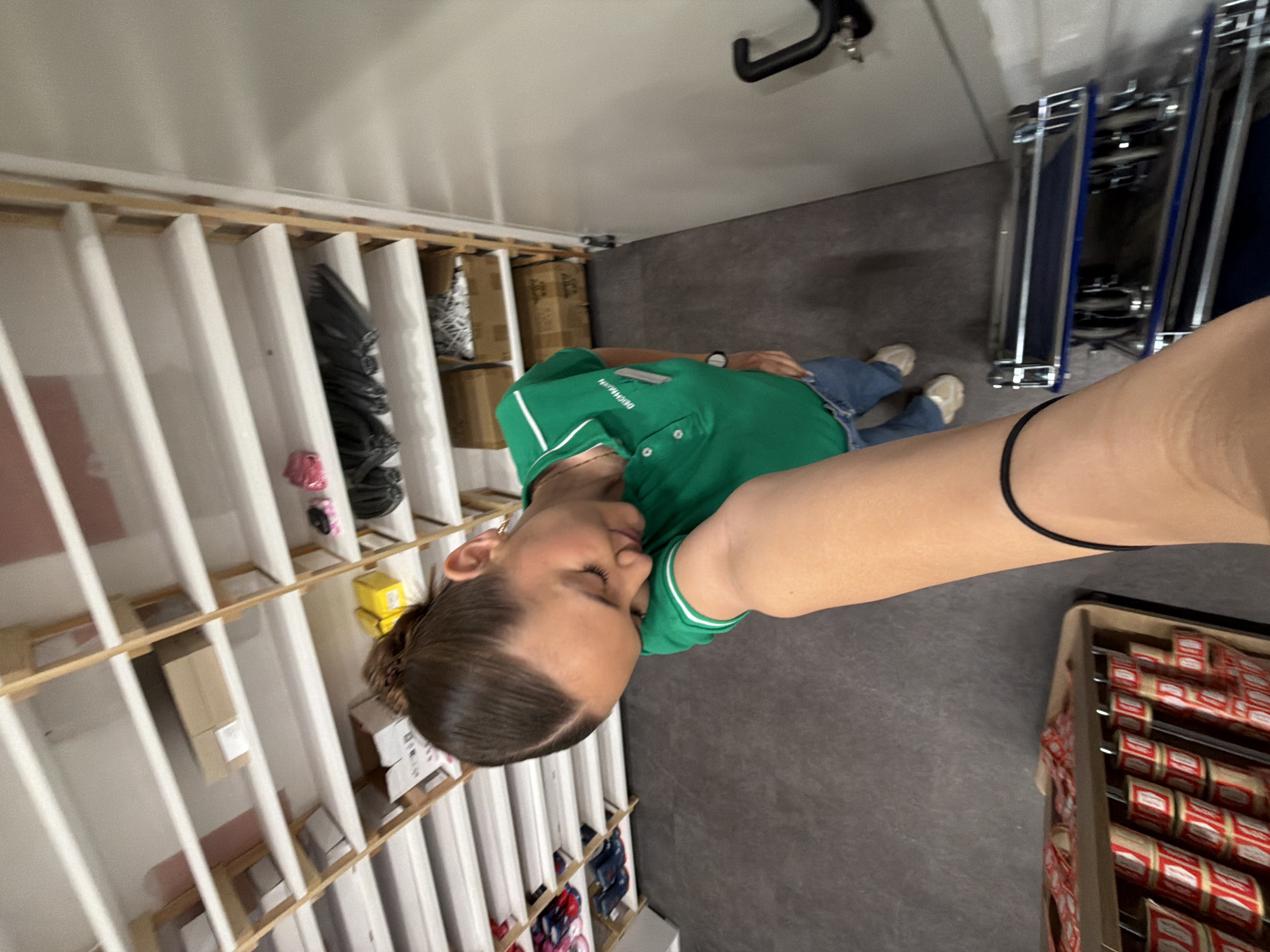 A first-person perspective photograph taken at an unusual angle in what appears to be a storage or retail setting. The subject, a fair-skinned individual wearing a green polo shirt with white trim and denim jeans, seems to be in a confined area.
To the left, there is a series of white shelves arranged vertically, filled with boxes and miscellaneous items. A metal rack on wheels stands on the right, partially filled with blue and silver components. There are also boxes of what appear to be labeled cans or jars in a container. The background is a uniform, grey wall or floor. The setting is likely a backroom or storage area within a retail establishment. The exact location in Sofia, Bulgaria cannot be determined from the image.