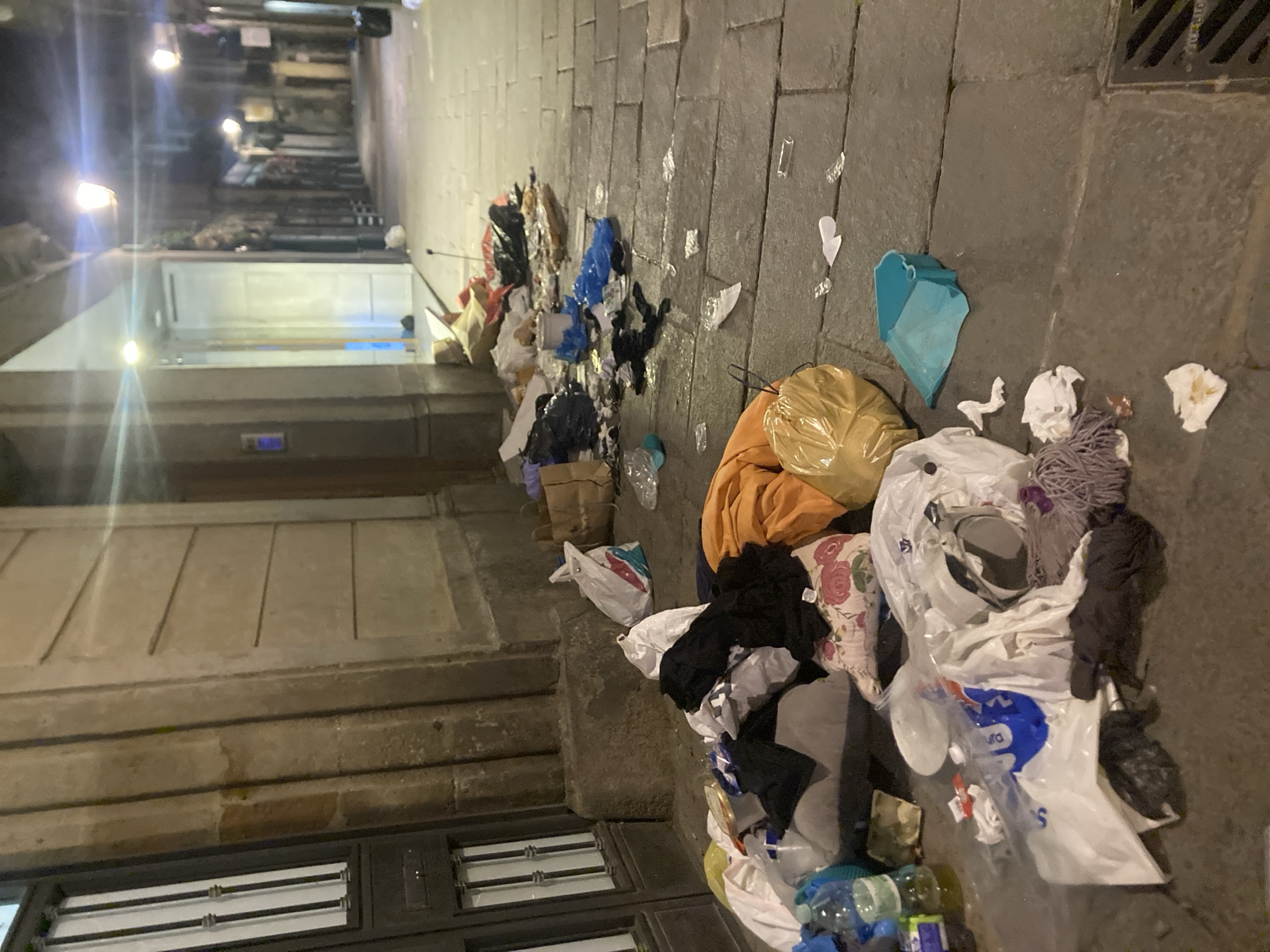 The image depicts a street scene in Barcelona, Spain, littered with trash and debris. Various types of refuse, including plastic bags, bottles, paper, and discarded clothing, are scattered along the cobblestone pavement and against the walls of buildings. The scene appears to be unkempt and unsanitary, with the accumulation of trash detracting from the urban environment. There are no people visible in the frame. The lighting suggests it is night.