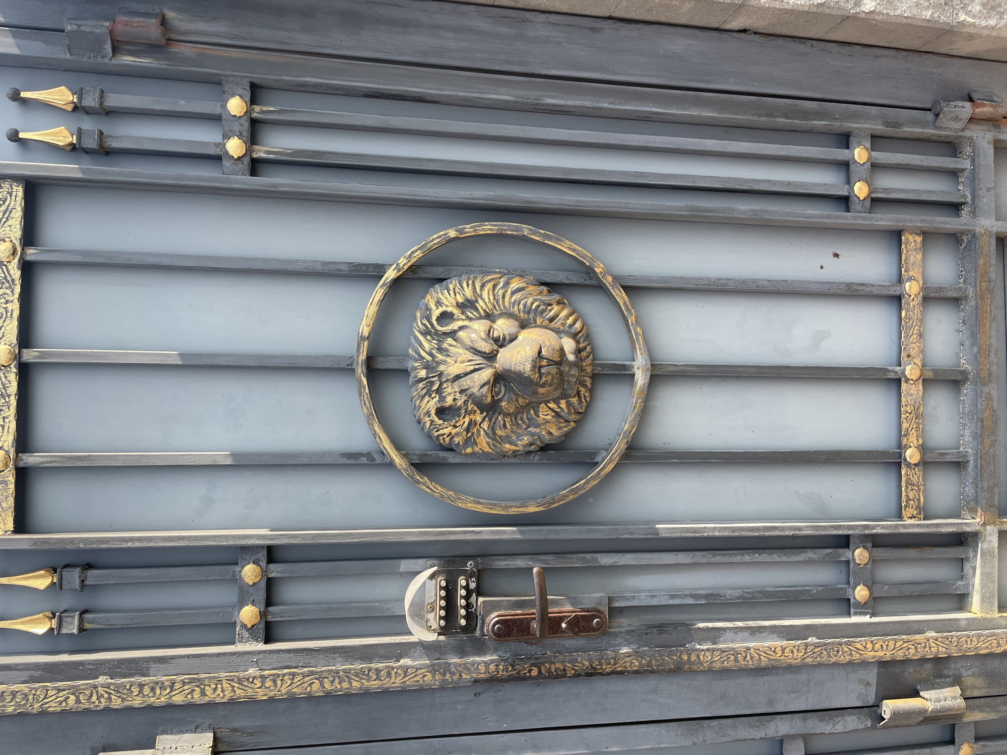 The image shows a close-up, eye-level shot of a gray metal gate with decorative gold accents. The gate features vertical bars with horizontal decorative elements, some of which are gold. In the center of the gate, a circular relief of a lion's head in gold and dark brown tones is prominently displayed. To the left of the lion's head, a dark brown metal door handle is attached to a vertical bar, and above it, a black electronic keypad with a clear plastic cover is mounted. The keypad has several rows of buttons, suggesting it is an access control system. The gate appears to be part of an entrance to a building or property. The lighting suggests it is daytime, with sunlight casting shadows. The overall impression is of a secure and somewhat ornate entryway. This gate is located in Almaty, Kazakhstan, specifically at the entrance of a private residence on Fursman Street.