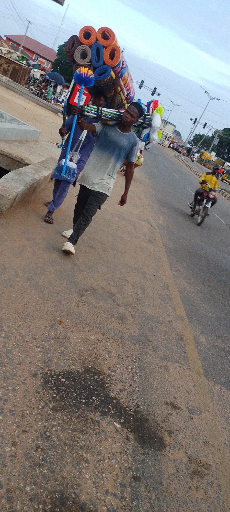 In Lokoja, Nigeria, a man walks along a paved street carrying a large bundle of rolled-up mats on his shoulder. He wears a blue-and-white t-shirt, black pants, and white shoes. Another person, behind the man, carries blue cleaning tools in a plastic bag. A motorcycle approaches on the road. The background shows traffic lights, buildings, and other pedestrians. The sky is overcast.