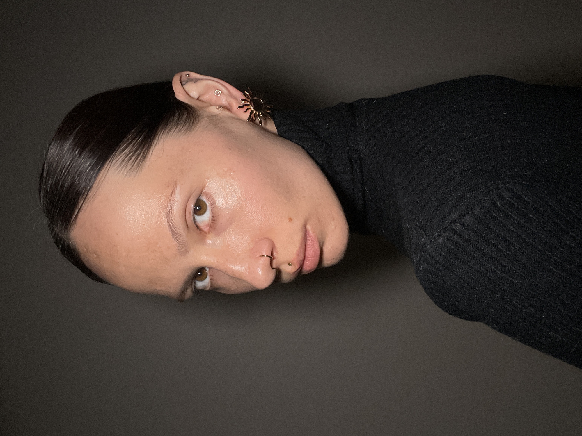 A close-up, rotated portrait shot features a young adult with dark, slicked-back hair, looking directly at the viewer. The image is oriented with the person's head tilted upwards, suggesting they might be lying down or the photo was taken from an unusual angle. The subject has clear skin, brown eyes, and several facial piercings, including a small gold hoop in their septum and a tiny stud on their upper lip, above the left corner of their mouth.
The visible ear is adorned with multiple small, shiny earrings, including a prominent gold-colored sunburst or spiky-shaped cuff or earring positioned on the outer rim of the ear, close to the helix. They are wearing a black, ribbed turtleneck sweater, the texture of which is visible, especially in the cropped images. The background is a plain, dark gray or black, providing a minimalist setting that keeps the focus entirely on the subject. The lighting appears soft and controlled, highlighting the contours of the face and the sheen of the hair. No specific activity is depicted beyond posing for the portrait. No text is visible in the image. The provided context indicates the location is Pervomaysky, Russia, though this is not visually evident.