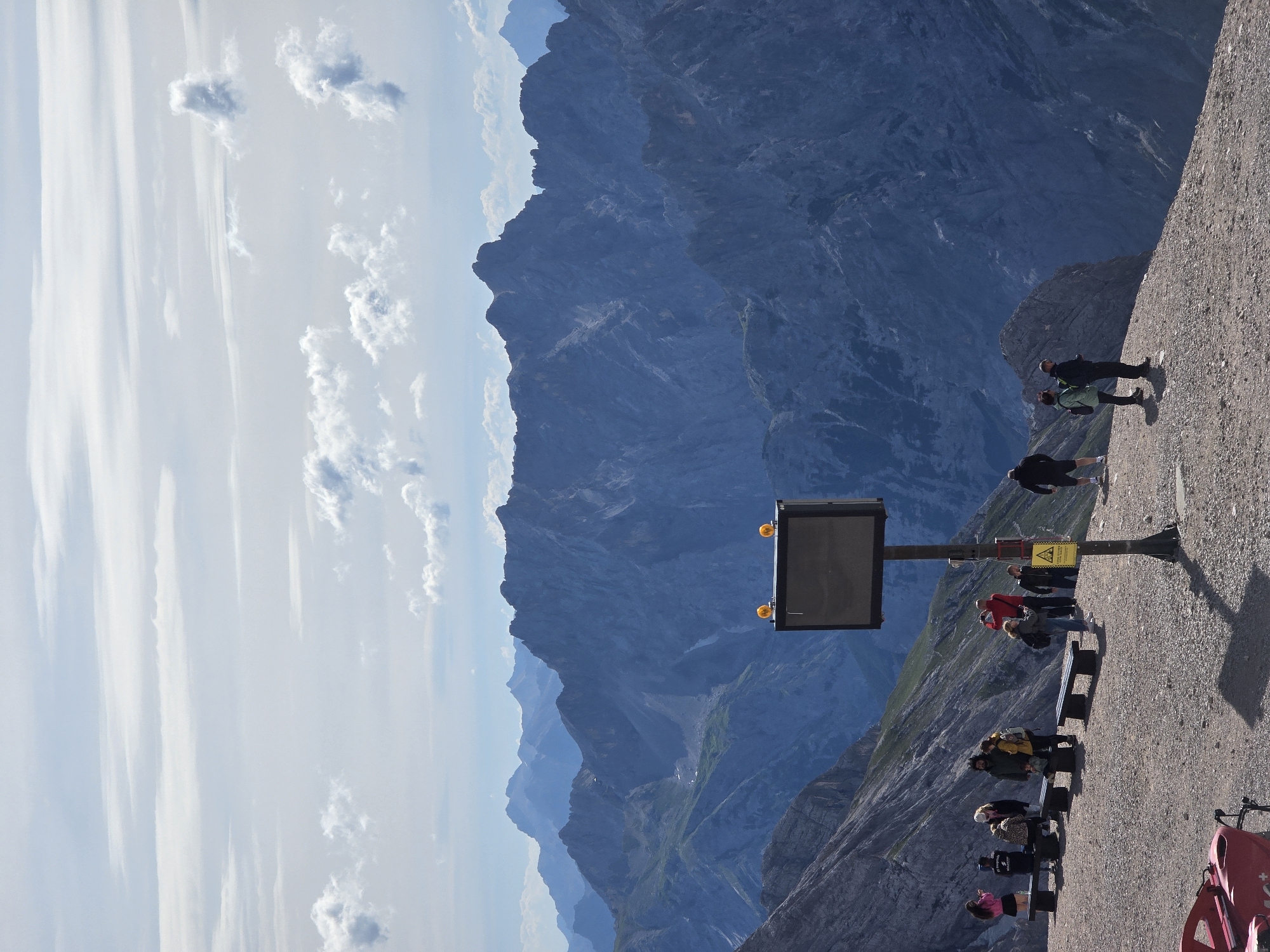 This is a photograph taken from the summit of the Zugspitze, Germany's highest peak, near Garmisch-Partenkirchen. The image captures a vast mountain landscape with multiple layers of rugged, dark blue peaks receding into the distance under a partly cloudy sky. In the foreground, a group of tourists are gathered on a gravelly plateau.
In the immediate foreground, on the left side, a red object, possibly part of a ski lift or equipment, is visible. To the right of this, a pole stands with a dark rectangular sign or screen mounted on top, equipped with two yellow warning lights. A yellow warning sign with a black symbol is attached to the pole lower down.
Several individuals are distributed across the plateau. On the left, near benches, a group of people are standing and sitting. Further to the right, a man in a red jacket is taking a photo with his phone. Other individuals are seen walking and standing, some with backpacks, observing the panoramic views. The terrain is rocky and uneven, typical of a high-altitude mountain environment. The overall impression is one of a popular tourist viewpoint on a clear, sunny day, offering breathtaking views of the Alpine mountain range.