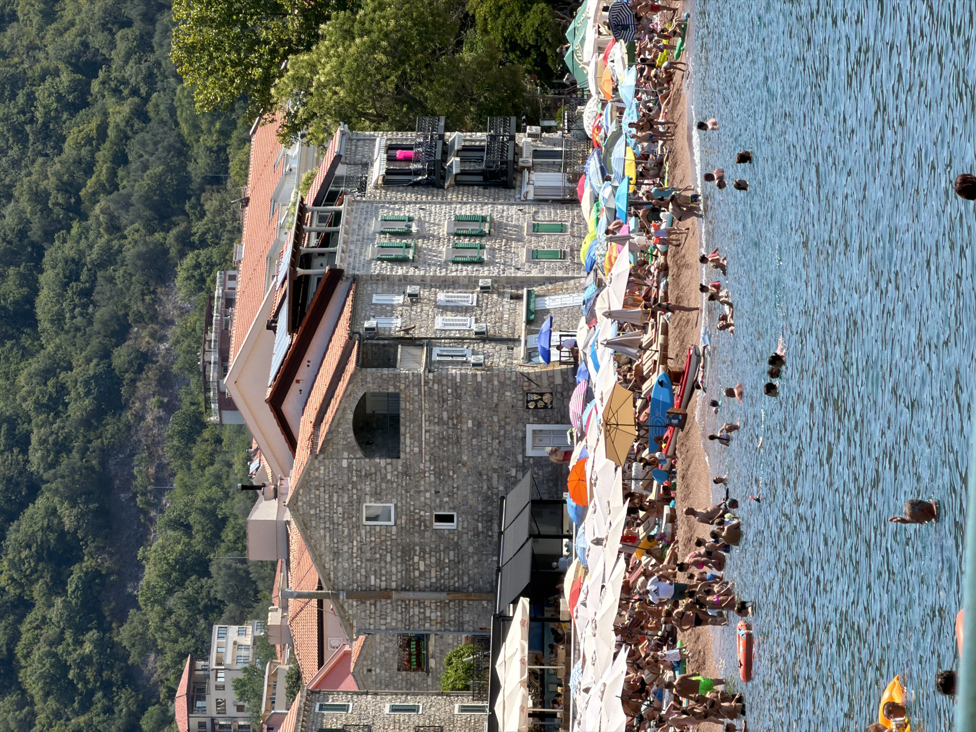 The image depicts a crowded beach scene in Petrovac centar, Montenegro, under a bright, sunny sky. In the foreground, the blue sea is filled with numerous people swimming, with some individuals wearing inflatable rings. The middle ground shows a bustling beach with a dense concentration of beachgoers lounging on chairs and towels under a multitude of colorful umbrellas. The beach is backed by a row of multi-story buildings constructed with stone and featuring red-tiled roofs. These buildings appear to be hotels or apartment complexes, some with balconies overlooking the beach. Behind the buildings, a lush green hillside densely covered with trees rises steeply. The overall impression is of a vibrant summer vacation spot with high tourist activity.