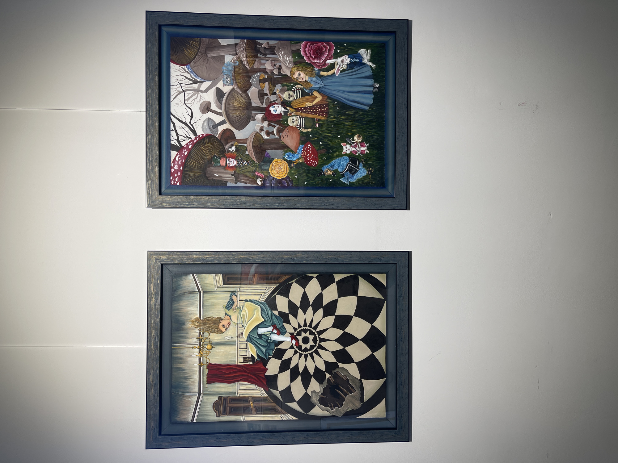 Two framed illustrations, seemingly depictions of "Alice in Wonderland," are hung on a white wall. The top frame contains an illustration of Alice surrounded by mushrooms, various characters, and objects associated with the story. The bottom frame depicts Alice falling through a floor with a black and white checkered design. The location is unknown and not discernible from the image. There are no people visible in the image except in the illustrations.