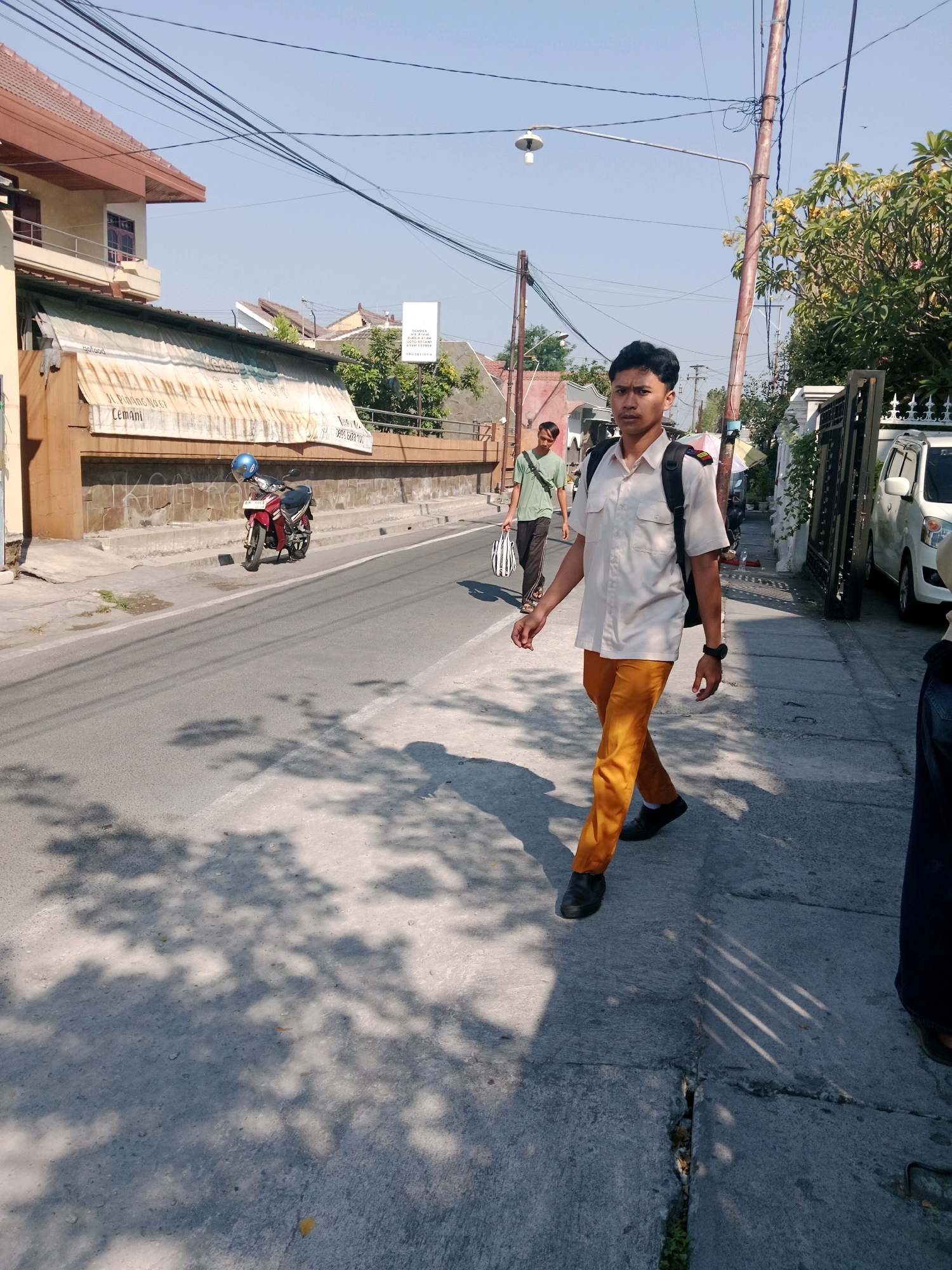 The image depicts a sunny street scene in Cemani, Indonesia. In the foreground, a young man wearing a light-colored, short-sleeved shirt and bright orange pants walks towards the camera on a concrete sidewalk. He has a black backpack on and is looking directly at the viewer. His right hand is extended slightly, and he wears a watch on his left wrist. Behind him, on the road, another young man in a green t-shirt and dark pants walks away from the camera, carrying a striped bag. A red motorcycle with a blue helmet on its handlebars is parked on the side of the road. Buildings with terracotta roofs line the street, and overhead power lines crisscross the sky. The environment appears to be a residential or commercial area, with trees and foliage visible along the sidewalk. The street itself is paved with asphalt, and the sidewalk is made of concrete. Shadows from trees are cast across the road and sidewalk, indicating a bright, clear day.