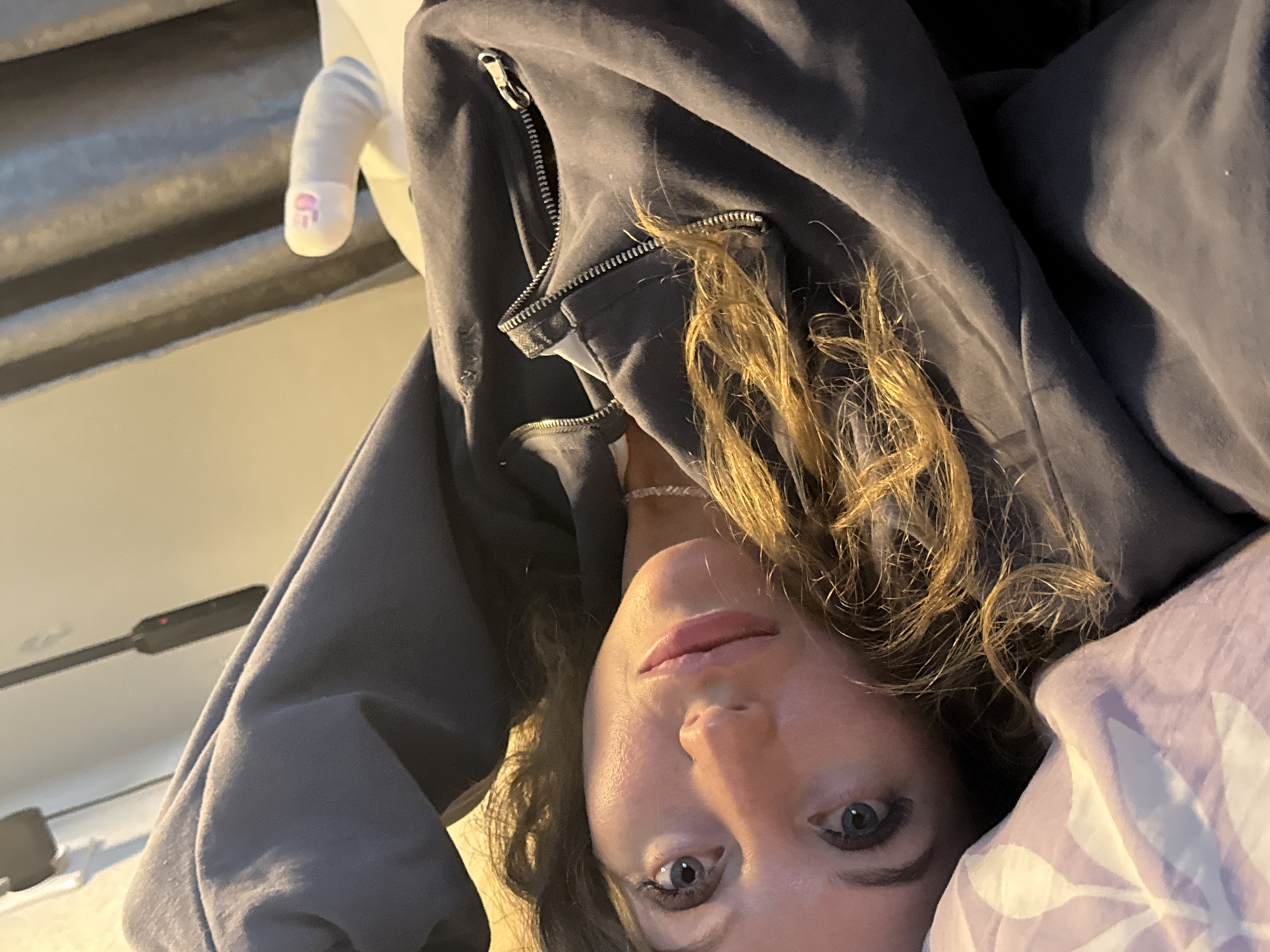 A woman with shoulder-length wavy brown hair and blue eyes is lying down, facing the camera. She is wearing a gray zip-up hoodie. Part of a white pillow with a floral pattern is visible to her right. To the left of her, a white plush toy with a pink design on its "finger" is visible. The background appears to be a room with a ribbed surface.