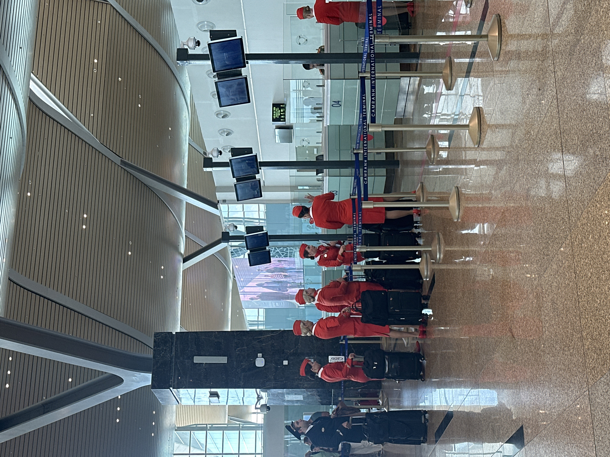 The image shows a group of airline staff at Cam Ranh International Airport in Bac Cam Ranh Ward, Vietnam. They are standing in a line at a check-in area, each with a suitcase. The airline staff are wearing red uniforms. The check-in area is marked by retractable barriers with "CAMRANH INTERNATIONAL TERMINAL" printed on the straps. Check-in counters are visible behind the staff, featuring monitors. The airport's interior is modern, with a curved wooden ceiling, large windows, and a polished floor.