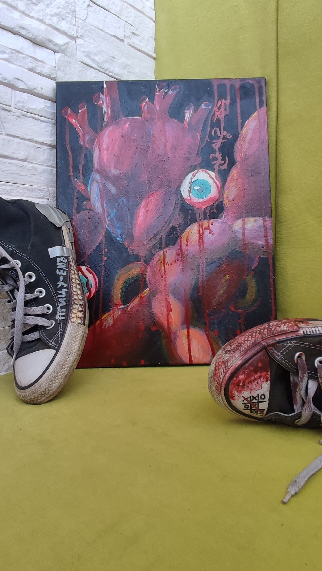 The image shows a canvas painting resting on a light green surface, flanked by two black canvas shoes. The painting depicts a stylized, fleshy, possibly monstrous figure with a large eye. Red paint drips are visible, suggesting blood or a visceral theme. The shoe to the left has Cyrillic script on its side. The shoe to the right has cryptic symbols. The background includes a white brick wall on the left and the edge of a green cushion or piece of furniture on the right. Location is not explicitly evident in the image, and therefore, Shelekhov, Russia, cannot be confirmed.