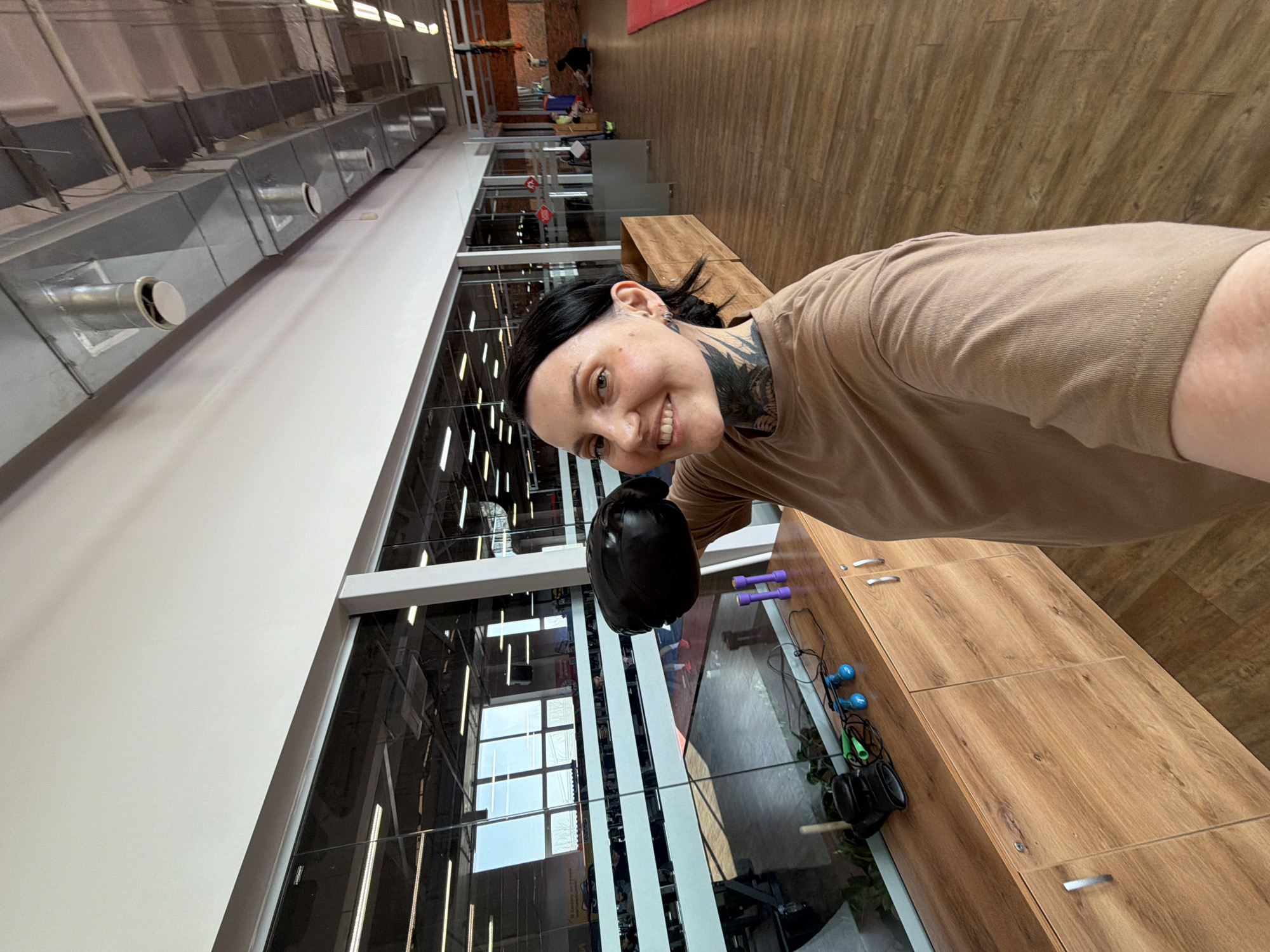 Here is a description of the image.
A selfie shows a woman with fair skin and tattoos wearing a tan t-shirt and holding a black object. Her dark hair is pulled back. The background is an indoor gym space. There are large windows behind her and a wooden cabinet to her right. The windows offer a view of the interior of the gym. Gym equipment can be seen in the reflection of the windows, including weights and machines. The location appears to be in Chaikovsky, Russia based on metadata.