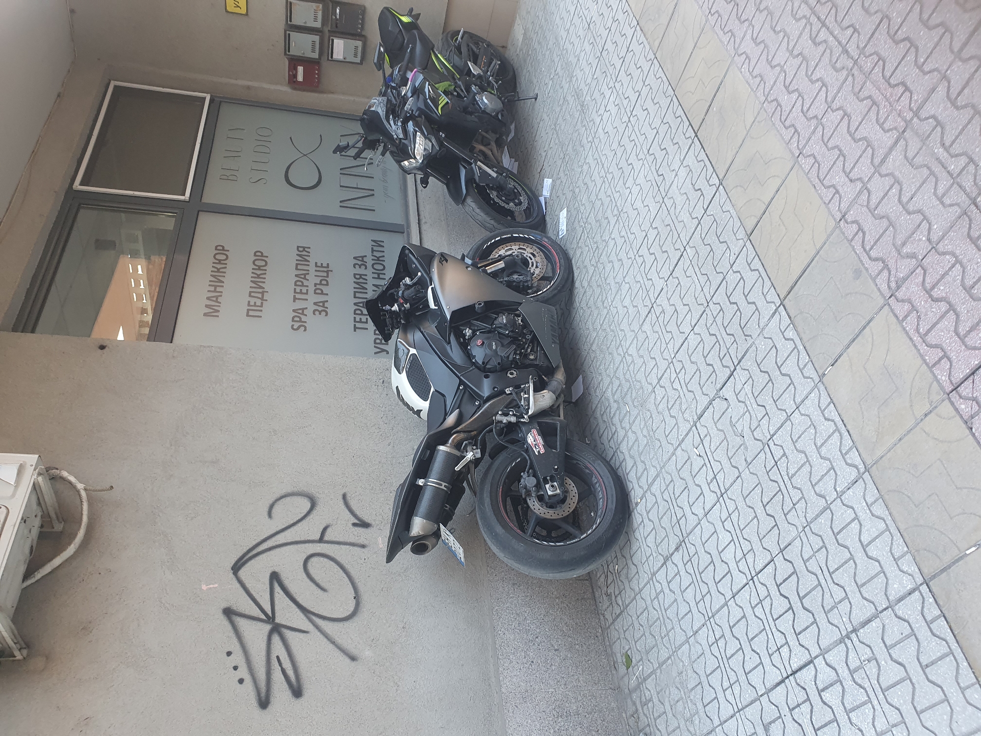 The image shows two motorcycles parked on a tiled sidewalk in front of a building in Plovdiv, Bulgaria. The building has a business called "Beauty Studio Infinity" written on the window in Bulgarian and English. The window also lists manicure, pedicure, and spa treatments. The wall next to the door has graffiti. Above the business entrance, there are mailboxes on the wall. The motorcycles appear to be parked legally. One is a black and green Kawasaki and the other is black and white. An air conditioning unit is located on the wall to the left of the door.