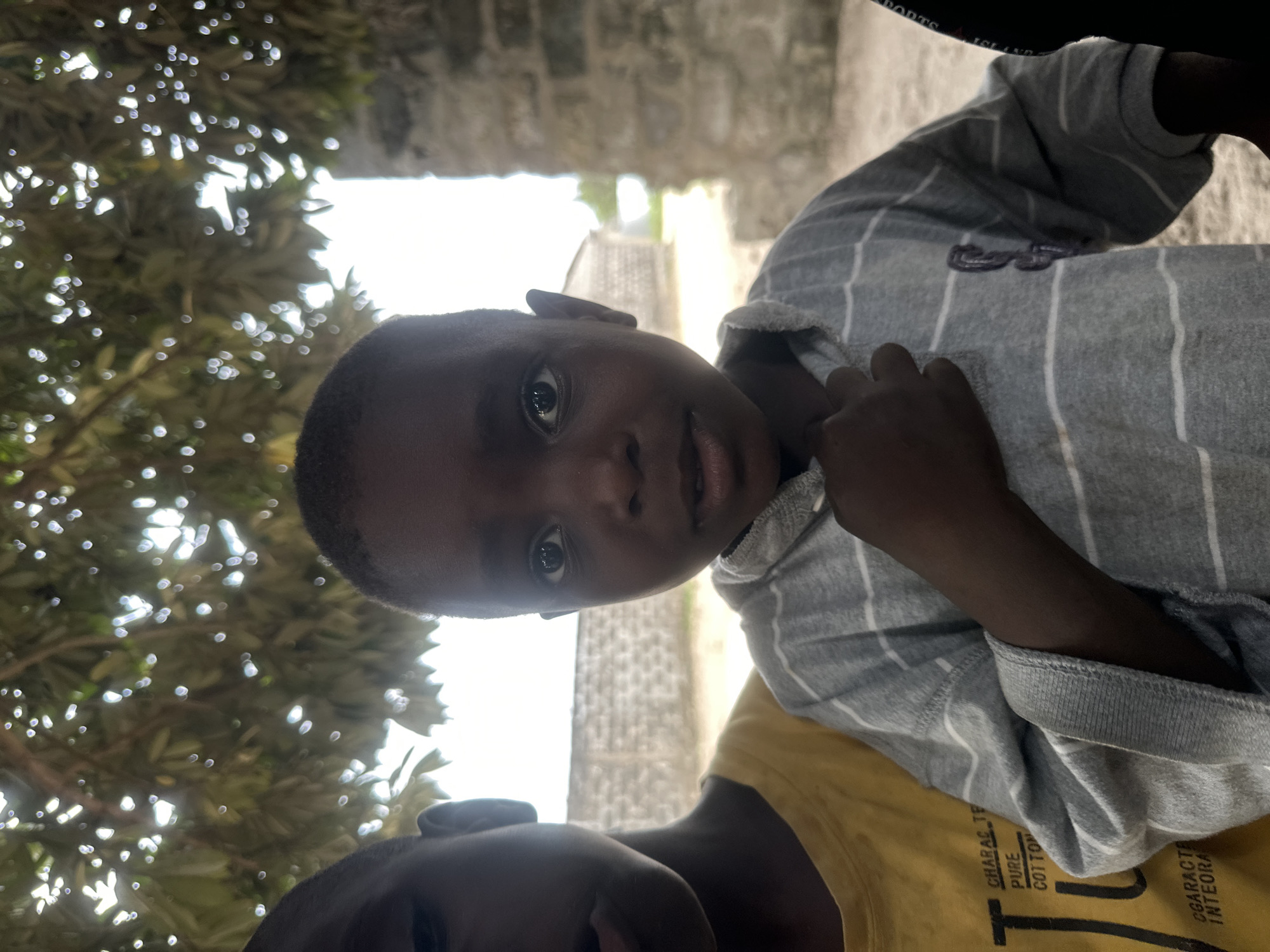 Two young children stand near each other outdoors. The child on the right, facing forward, is in focus and appears to be the primary subject. This child has dark skin and is wearing a grey and white horizontally striped long-sleeved shirt. Their right hand is raised to their chest, near their collarbone. The child on the left is partially visible on the left side of the frame, facing left. This child is wearing a yellow t-shirt with black text that appears to read "CHARAC TE PURE COTTON" and "CARACTE INTEGRA". The background shows a beige, sandy ground, a low grey brick wall with a repeating pattern, and some blurred buildings in the distance. Above the children, foliage from a tree casts dappled shadows. The scene is outdoors, likely in a village or rural area, given the simple structures and natural elements. The image has a shallow depth of field, with the background out of focus. The overall lighting suggests it is daytime.