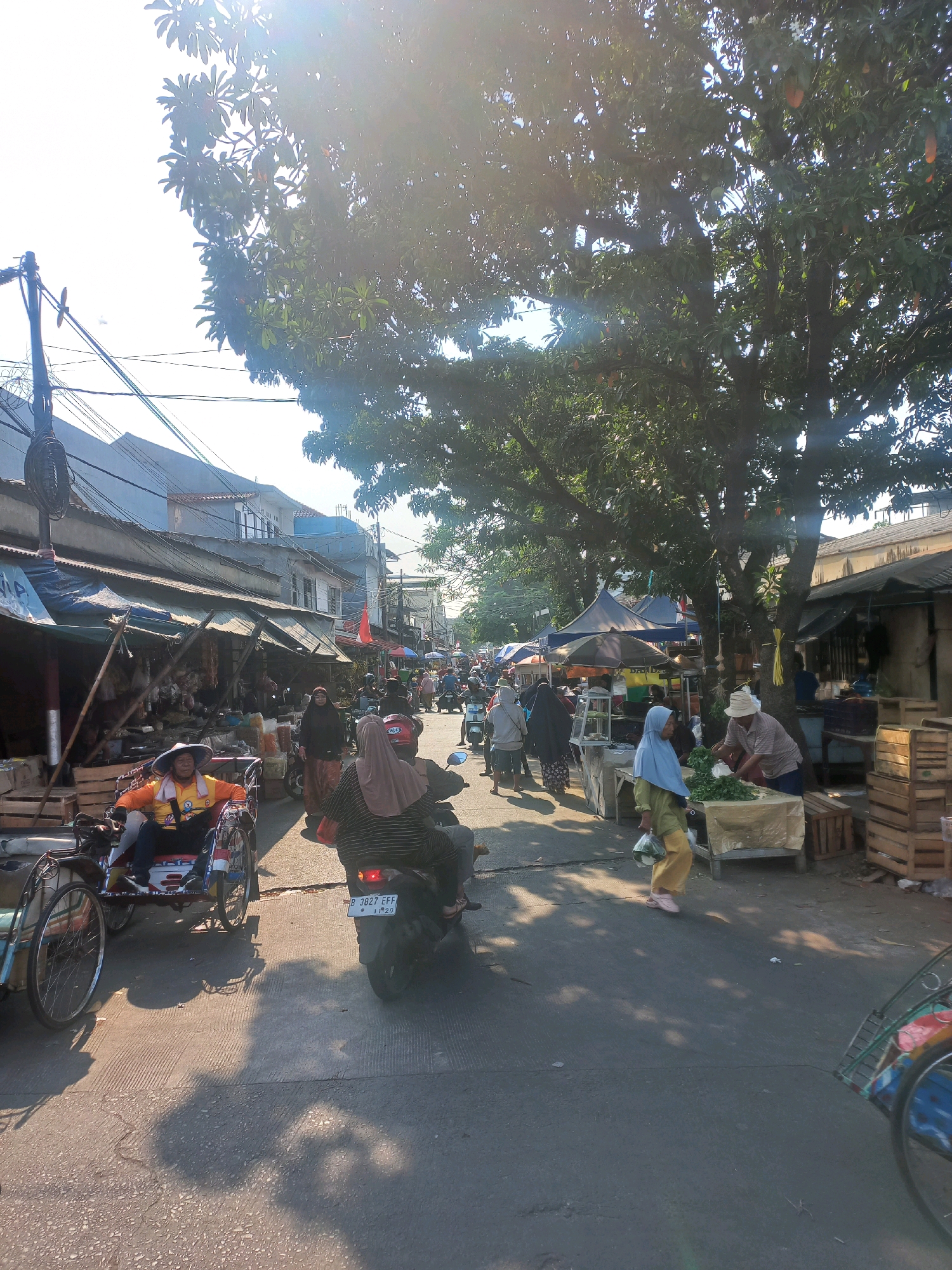The image depicts a bustling street market in Depok, Indonesia, likely on a sunny day given the strong shadows and lens flare. The scene is filled with activity, with numerous people engaged in various tasks. In the foreground, a motorized pedicab driver, wearing an orange shirt and yellow vest, is seated, looking ahead. Behind him, a person on a motorcycle, wearing a headscarf and patterned top, is visible from the back. Several other individuals are seen walking and riding motorcycles along the street, which is lined with market stalls. These stalls feature awnings and are filled with goods, including produce. Some stalls display crates of vegetables, and a vendor is seen attending to a customer. Overhead, large trees cast shadows on the street, contributing to the dappled light. Power lines and utility poles are present along the side of the street, indicating an urban setting. The overall impression is that of a vibrant, active marketplace in Indonesia.