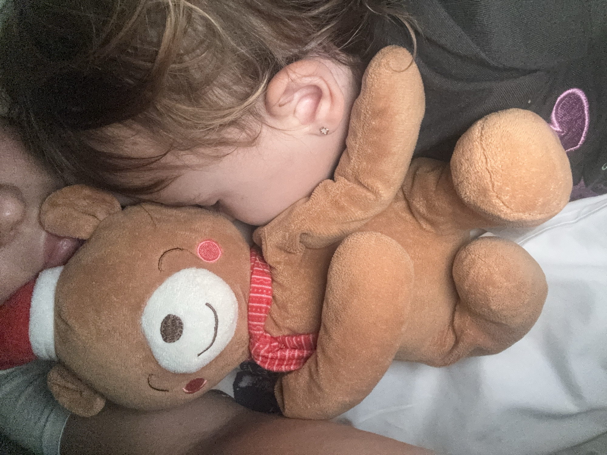 A young child is sleeping on a white sheet next to a brown plush reindeer toy with a red and white scarf. The child has fair skin, light brown hair, and a visible ear with a small star-shaped earring. The child is positioned to the left of the reindeer, with their face resting close to the toy. A person's face is partially visible to the left of the child, possibly lying next to them. The background includes a dark fabric with a purple embroidered detail.