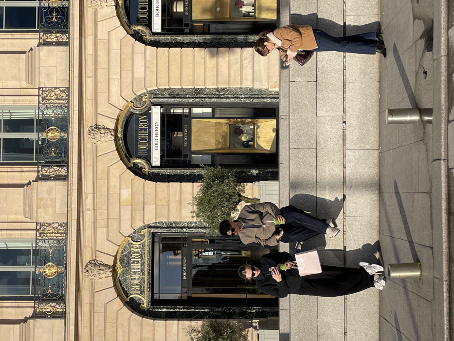 The image captures a street scene in front of the Boucheron boutique at 26 Place Vendôme, Paris, France, under bright sunlight. The building features light-colored stone facade with classical architecture, including multiple arched entrances adorned with gold "BOUCHERON" lettering and detailed mascaron carvings above. Ornate black wrought-iron balconies with gold accents are visible on the upper floors.
In the foreground, a multi-tiered gray stone ledge separates the building from the street level. Three individuals are present. On the left, a female stands facing her mobile phone, dressed in a black long-sleeved top, black wide-leg trousers, white sneakers, and dark sunglasses. She holds a green drink in her right hand and a light pink shopping bag in her left. Beside her, on the stone ledge, a male sits facing his mobile phone. He wears a light brown jacket, dark trousers, light-colored sneakers, and dark sunglasses, holding a similar green drink. To the right, a second female stands, also looking at her mobile phone. She is wearing a light brown jacket with a cream faux-fur collar, dark blue jeans, and black boots, carrying a dark reddish-brown shoulder bag.
Two small olive trees in planters are positioned on the stone ledge behind the individuals. Two silver cylindrical bollards are visible on the sunlit paved street in the lower foreground. Long, distinct shadows are cast across the pavement, indicating strong, direct sunlight from the left side of the frame.