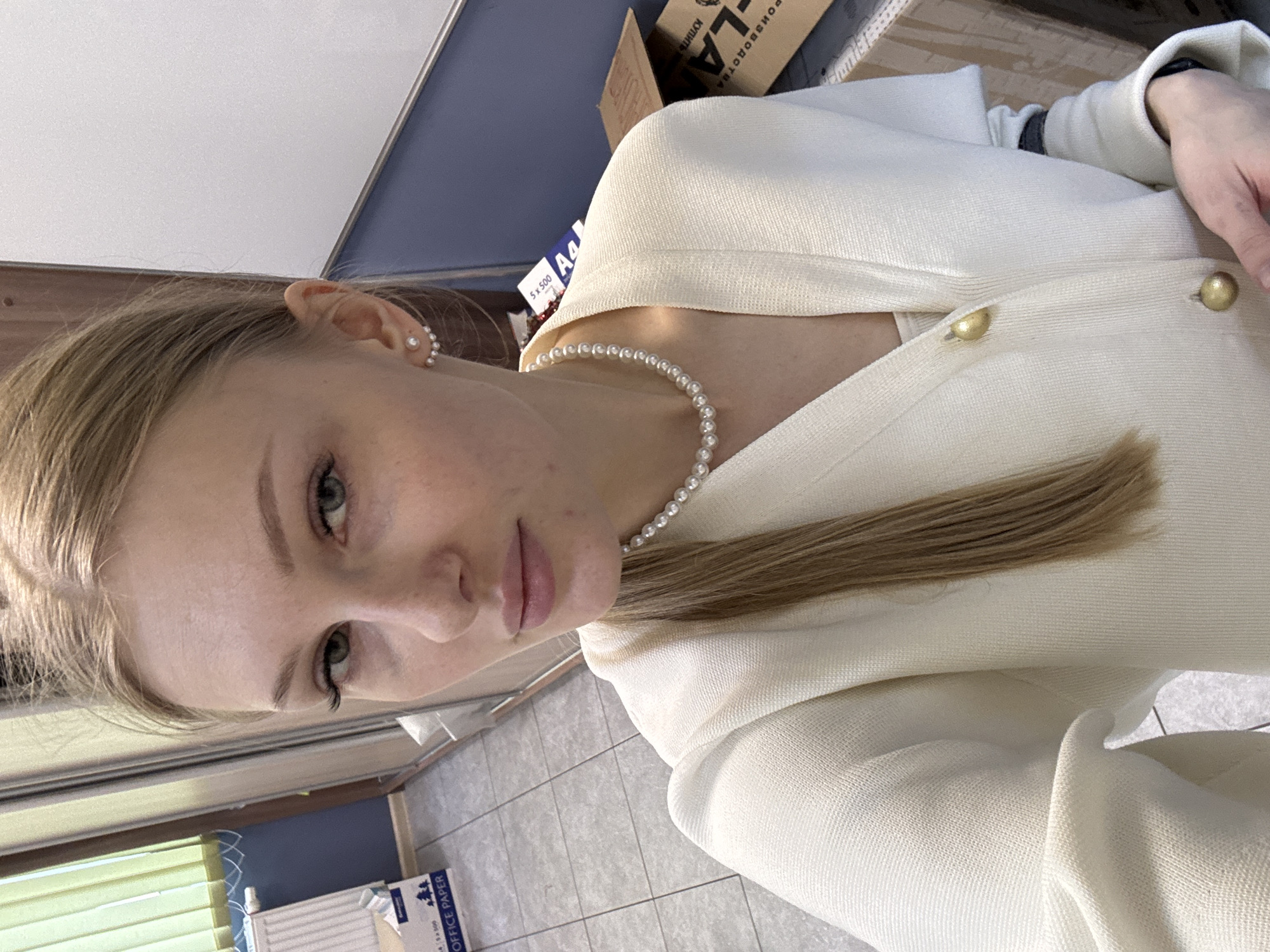 A young blonde woman is facing the camera, wearing a pearl necklace and a white jacket with gold buttons. She also has pearl earrings. She is indoors. Behind her are office supplies including a box labeled "LA-KLA," and a pack of paper labeled "A4". The location appears to be Perm, Russia, based on the presence of Cyrillic text.