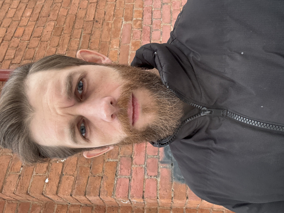 A male subject, appearing to be in his 30s to 40s, is captured in a close-up outdoor portrait. He has light brown hair, blue eyes, and a full light brown beard. The subject wears a black puffer jacket, zipped up, with a visible black zipper. Minor white specks, consistent with snowfall or dust, are visible on the jacket. He looks directly at the camera with a neutral expression.
The background consists of a weathered reddish-orange brick wall, exhibiting variations in brick color and condition. The wall forms an exterior corner on the left side of the frame. A partially obscured dark gray/black graffiti marking is visible on the lower section of the brick wall behind the subject's jacket. In the upper right background, a dark red architectural element, possibly a pipe or structural beam, is present. No specific landmarks or unique architectural details are present to identify a precise location within the city of Saint Petersburg, Russia.
