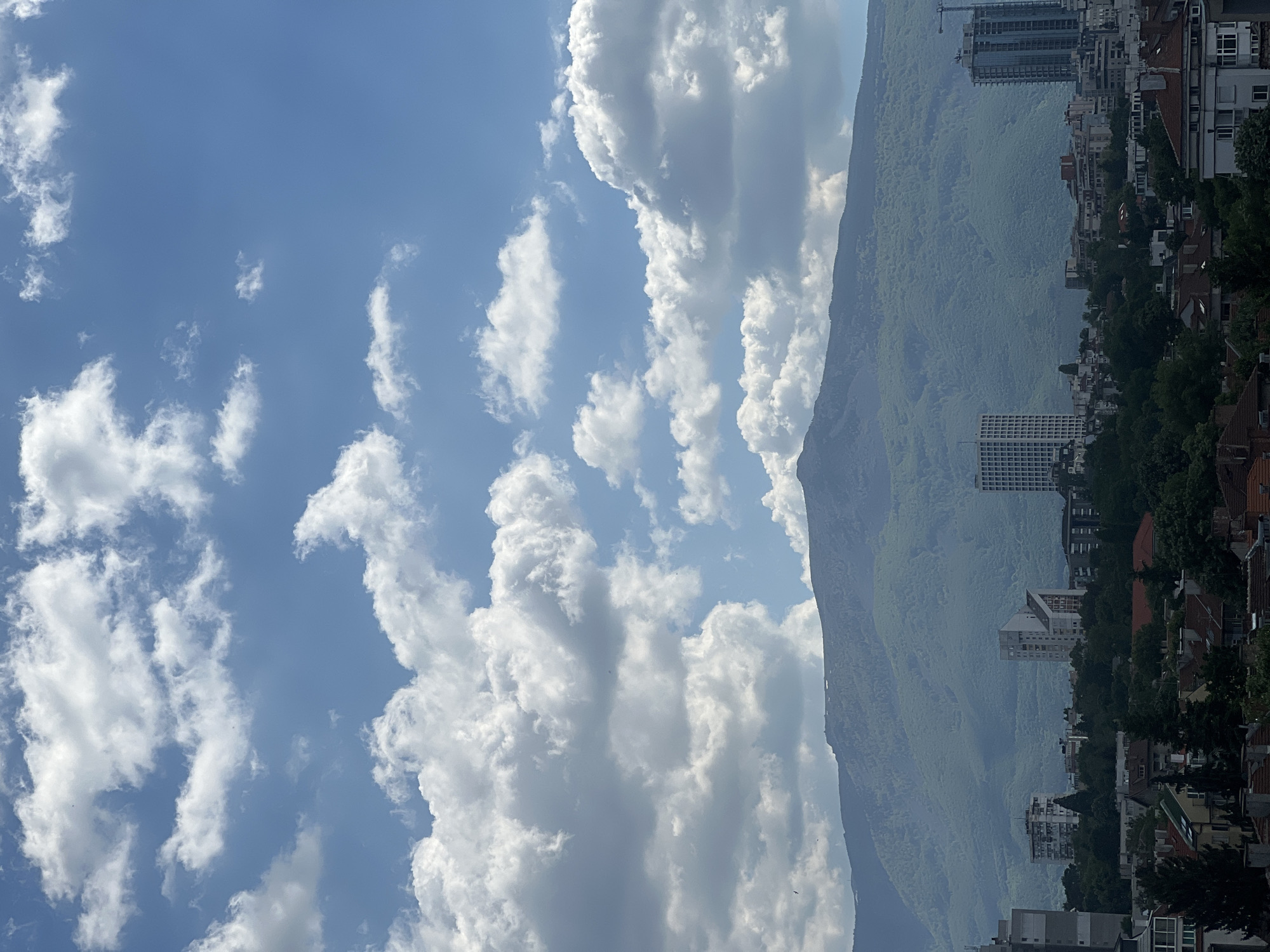 The image shows a view of Sofia, Bulgaria, with the Vitosha mountain range in the background. The sky is blue with scattered cumulus clouds. To the right of the mountain, there are residential buildings and some taller office buildings. Some construction cranes are visible amongst the buildings. The foreground contains rooftops and trees. The general impression is a clear summer day.