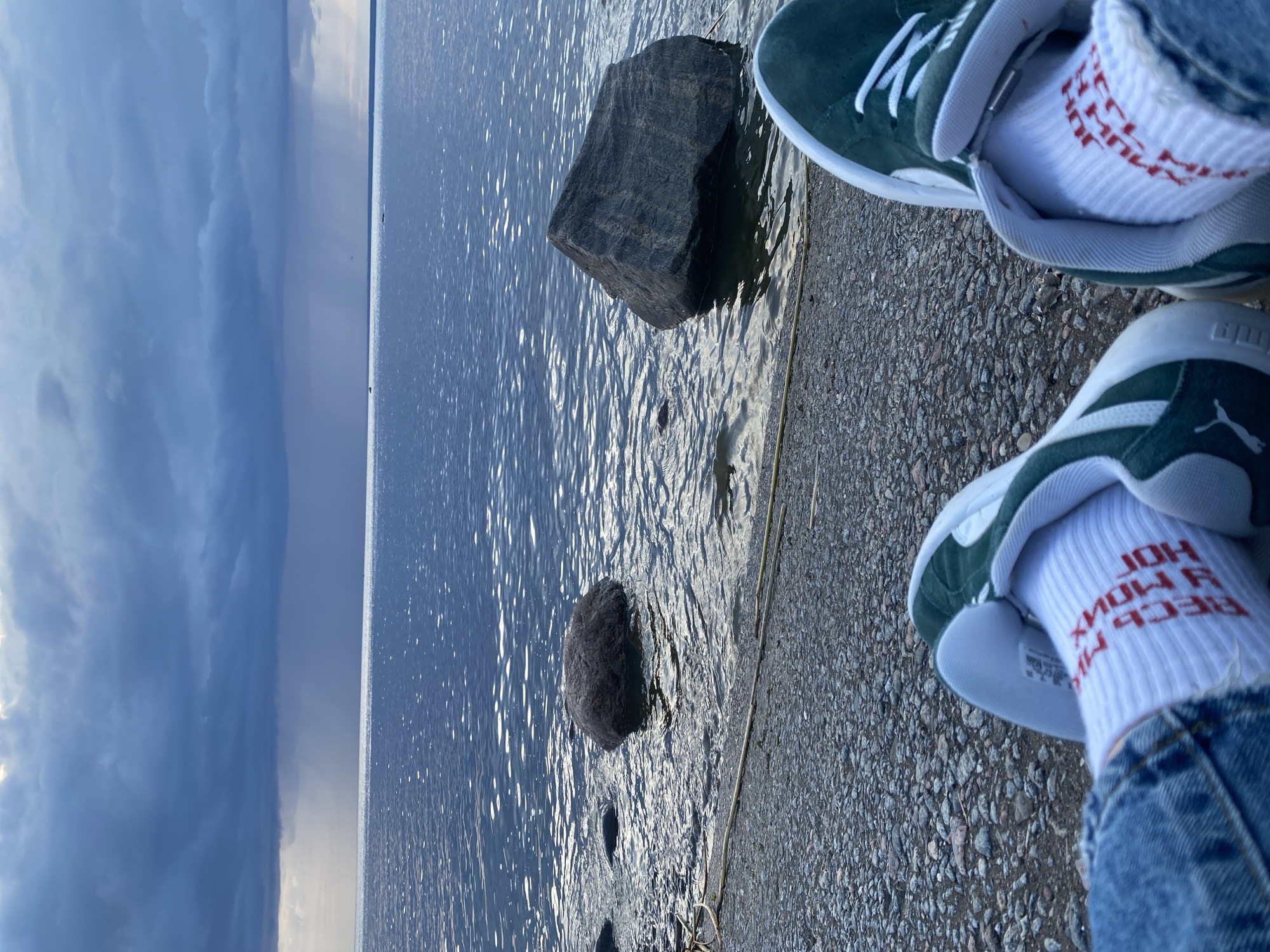A first-person perspective shot shows a person sitting by a body of water. The person is wearing blue jeans and green and white sneakers. White socks with red lettering are visible. The water is calm and reflects the sky. In the distance, there is a dark, cloudy sky with rain visible falling. Two large rocks are partially submerged in the water near the shore. The shore itself is covered in pebbles. The scene appears to be a waterfront location in Sestroretsk, Russia, likely during inclement weather.