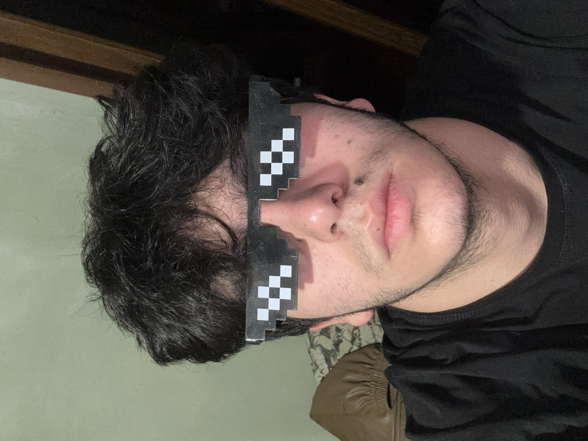 A close-up, tilted portrait shot depicts a person wearing "deal with it" pixelated sunglasses. The person has dark, curly hair and a light skin tone with stubble. They are wearing a black shirt. The background includes a green wall and a dark wooden trim. A piece of patterned fabric is partially visible near the bottom of the frame. No location information or specific details indicate Yerevan, Armenia.