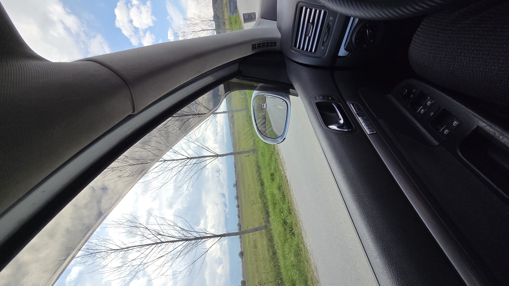 The image captures a view from inside a car, looking outwards. The perspective suggests the camera is held vertically, resulting in a landscape orientation where the car's interior is on the right and the exterior view is on the left.
Inside the car, the textured fabric of the A-pillar is prominent on the left, curving towards the top of the frame. To its right, parts of the dashboard and a silver air vent are visible. Further right, a portion of the steering wheel with a textured grip can be seen. The car's side mirror reflects a segment of the road and a distant landscape, with a small rectangular object (possibly another vehicle or an indicator) visible within the mirror's reflection. Below the mirror, the car's door panel is visible, featuring a chrome door handle, controls for power windows and side mirrors, and a metallic trim piece with the embossed text "LIMITED EDITION". The car's seat fabric is partially visible in the upper right corner.
Outside the car, a paved road runs horizontally across the middle of the frame (if viewed upright), bordered by a vibrant green field. Several bare, deciduous trees are spaced out along the side of the road, casting long shadows. The sky above is bright blue with scattered white clouds. The overall setting appears to be a rural or semi-rural area on a clear day, likely in late autumn, winter, or early spring due to the leafless trees. The car appears to be either stationary or driving alongside the road.