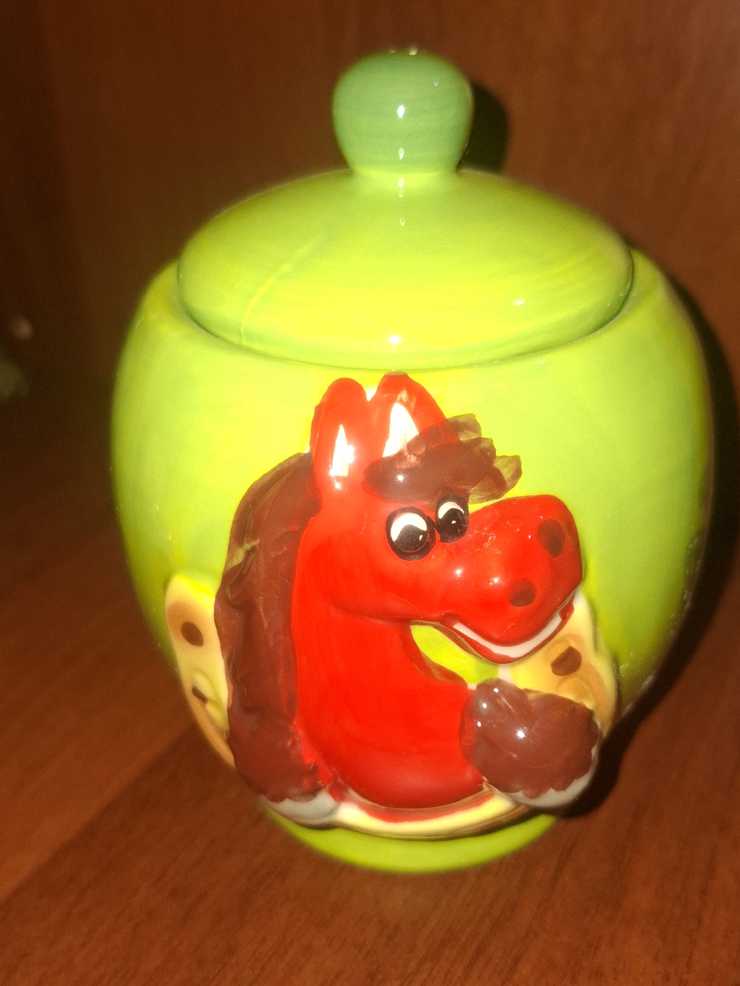 A close-up, eye-level shot shows a bright lime-green ceramic pot with a lid, which appears to be a sugar bowl or similar container. The pot is decorated with a raised, cartoonish red horse head. The horse has large white eyes with black pupils, a dark brown mane and forelock, and brown accents on its cheeks. The horse's mouth is open, revealing a white inner lip. There are also lighter yellow and brown elements visible on the sides of the pot, suggesting other decorative features, possibly representing cheese or similar textures, that might frame the horse. The pot is placed on a brown wooden surface, likely a table, with diffused lighting creating subtle shadows. The background is a dark, blurry brown. The image does not contain any information about the city of Almaty, Kazakhstan.