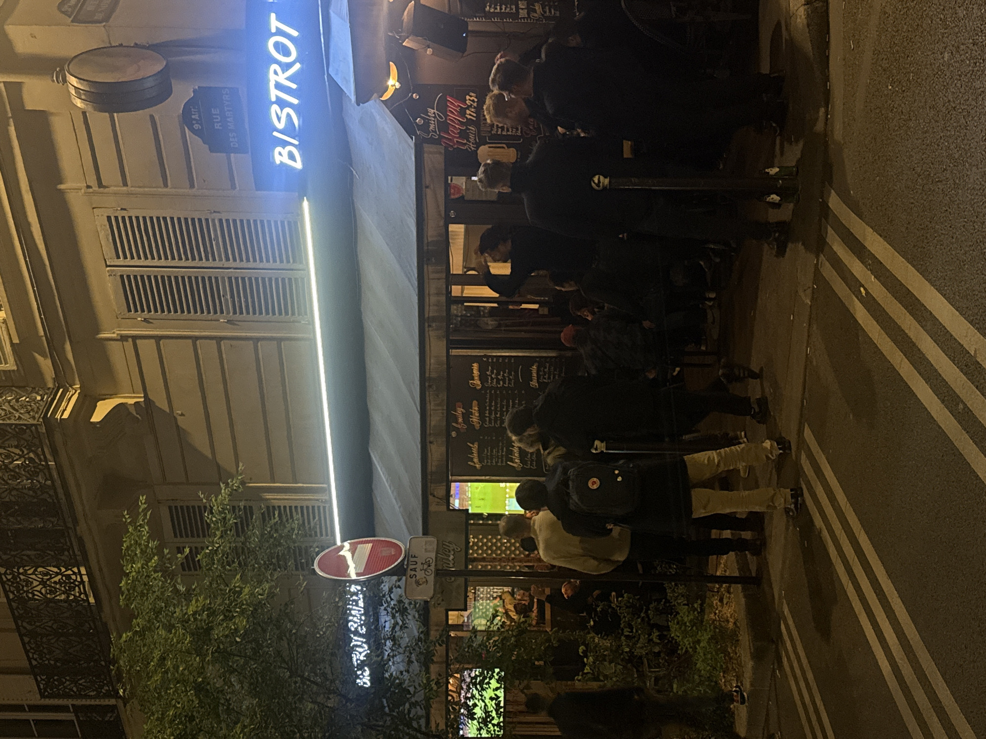 The image depicts an urban scene at night in Paris, France. People are gathered outside of a bistro, a sign indicates the location as "Rue des Martyrs, 9th Arrondissement" (9. Arr). The bistro, named "BISTROT" (with some additional signage indicating "BE TROT ZALLA"), appears to have a television screen visible from the street, attracting a crowd watching something. The street has striped markings. Vegetation is present. The scene is oriented at an angle, presumably to capture both the establishment and street detail. The scene is dimly lit with ambient lighting from the bistro and other sources.