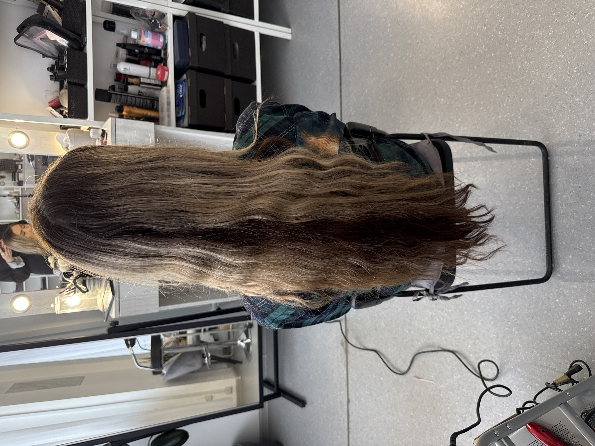A waist-up shot from behind shows a person sitting in a black chair in a brightly lit room. The person has long, wavy brown hair with blonde highlights, and is wearing a green and blue plaid shirt. The person is facing a vanity with a lighted mirror. A shelf with various makeup products and supplies is behind the vanity. The background is a speckled grey floor. The location cannot be determined to be Izhevsk, Russia from the visual information.