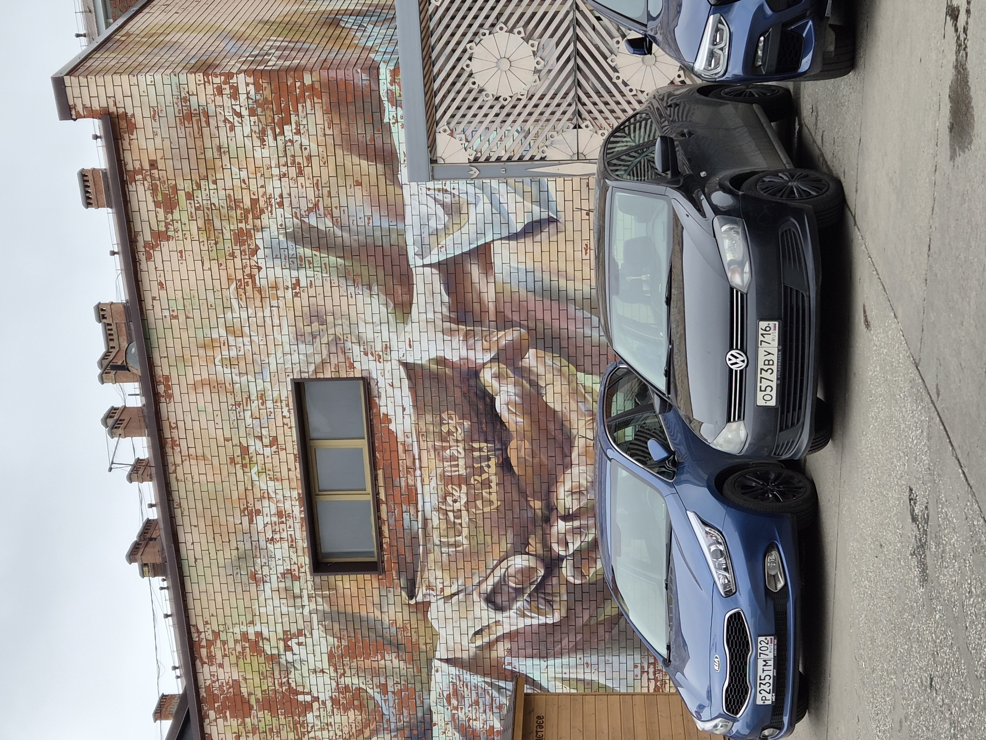 This is a medium shot, eye-level, outdoor, daytime photograph of a brick building facade with a mural and several parked cars in front. The building is made of red and beige bricks. A large mural is painted on the upper portion of the wall, depicting what appears to be hands holding a cup. The mural has a faded, abstract quality with warm tones. A horizontal, rectangular window with two panes and a brown frame is visible above the mural. Chimneys with decorative tops protrude from the roofline. To the right of the mural is a section of a decorative metal gate or screen with a geometric pattern.
In the foreground, three cars are parked on a paved surface. The leftmost car is a blue Kia hatchback with black wheels and a Russian license plate that reads "P235TM 702". The middle car is a dark grey Volkswagen sedan with black wheels and a Russian license plate that reads "O573BY 716". The rightmost car is a blue BMW SUV, only partially visible. The pavement has some cracks and dark stains. A small wooden structure with a sloped roof is visible to the left of the blue Kia. The sky is overcast and grey. The scene appears to be in an urban or industrial area. There are no people or animals visible in the image.