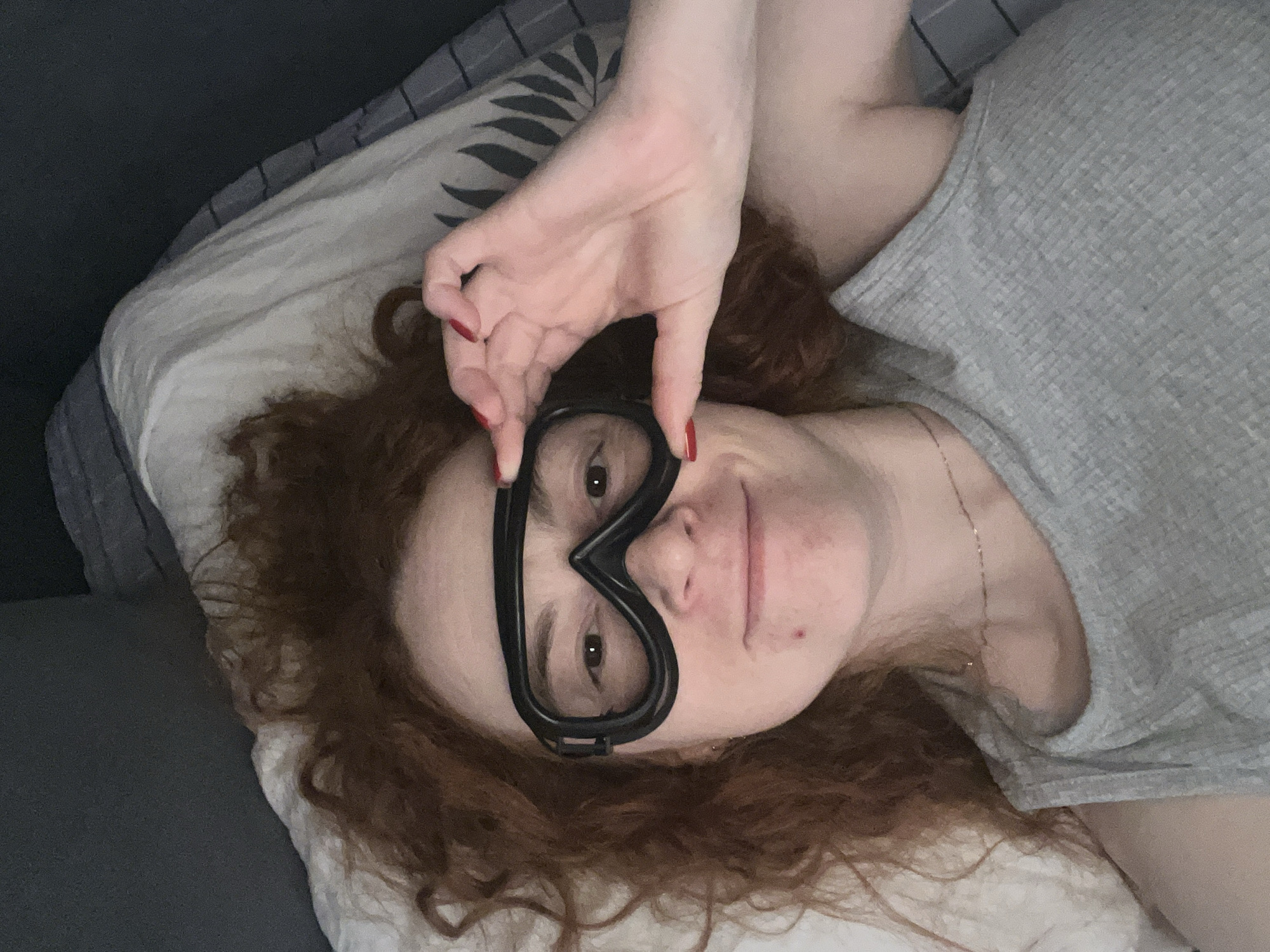 An overhead shot captures a person with auburn, curly hair lying on what appears to be a bed or sofa. The individual is looking directly at the camera with a subtle smile. Their eyes are partially covered by a pair of black, ski-style goggles or a mask, which they are holding in place with both hands. Their fingers, adorned with red nail polish, are visible near the top and bottom edges of the goggles.
The person is wearing a light grey, ribbed t-shirt, and a thin, delicate gold-colored necklace is visible around their neck. They are resting on a pillow with a patterned case featuring dark grey stripes and a faint, darker leaf motif. To the left, a solid dark grey blanket or bedding is visible. The setting is indoors, suggesting a relaxed environment like a bedroom. The lighting is soft and even, characteristic of indoor ambient light. No text is visible in the image.