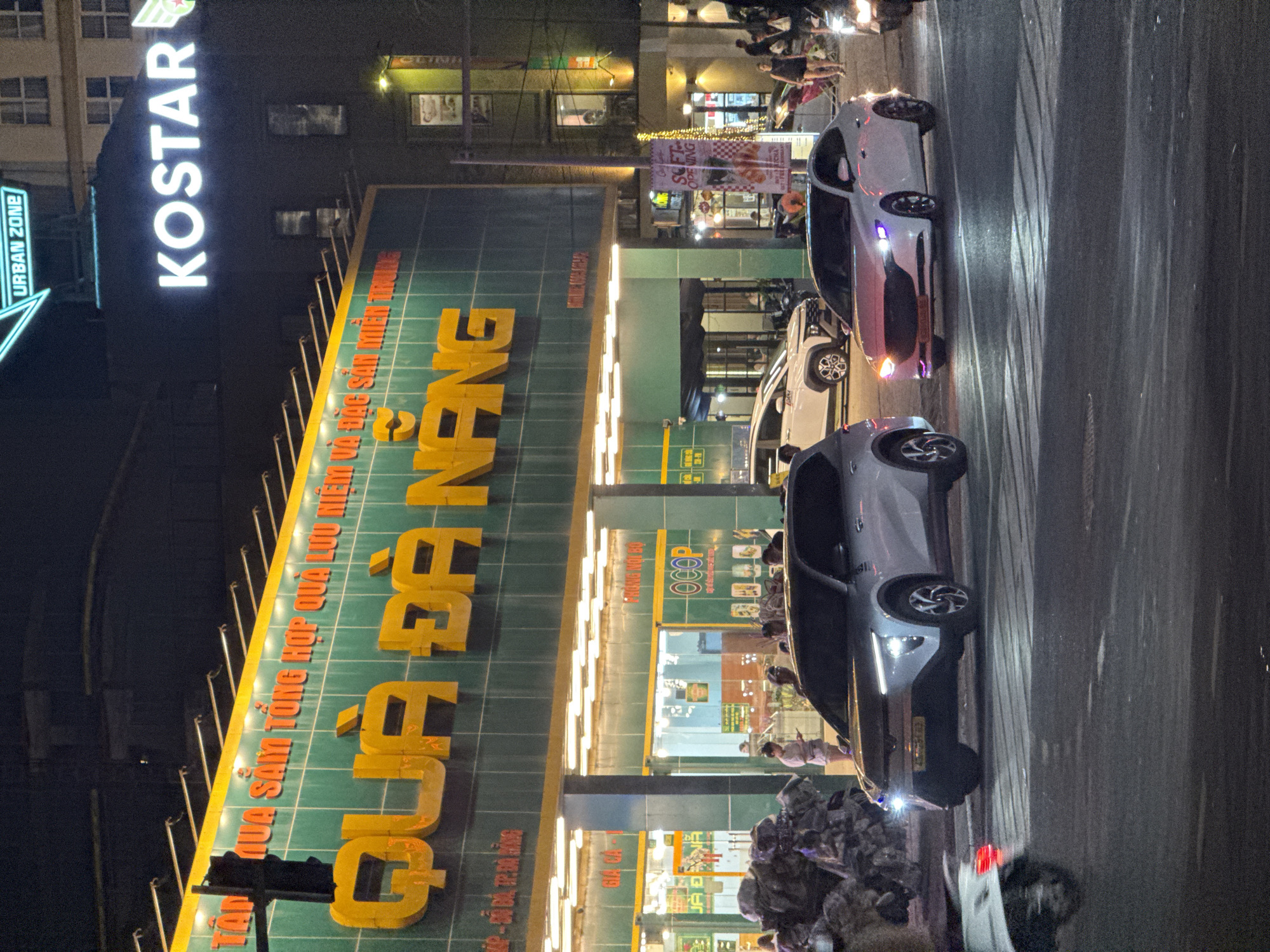 This nighttime street scene in Đà Nẵng, Vietnam, features a bustling commercial area dominated by a brightly lit multi-story building. The building's facade displays prominent signage in Vietnamese. Large yellow letters on a green background read "QUÀ ĐÀ NẴNG," indicating a gift or souvenir shop. Above this, smaller text states "QUÀ SẴM TỔNG HỢP QUÀ LƯU NIỆM VÀ ĐẶC SẢN MIỀN TRUNG," translating to "General Gifts, Souvenirs and Central Vietnamese Specialties." A large, vertical neon sign on the upper part of the building spells "KOSTAR" in white letters, with a smaller "URBAN ZONE" sign nearby.
On the street, several cars are visible, some parked and others in motion, their headlights and taillights glowing against the dark road. Two modern cars, one light-colored and one darker, are prominent in the foreground and mid-ground. The road surface appears to reflect the ambient light, suggesting it might be wet or simply highly reflective. People are seen on the sidewalks and near the storefronts, indicating a lively urban environment. A banner announcing "SOFT OPENING" is visible in the background, along with an "OCOP" logo and other smaller Vietnamese text like "TRANG NỘI BỘ" and "GIA CẢ." The overall scene portrays a vibrant, active commercial district during the evening hours.