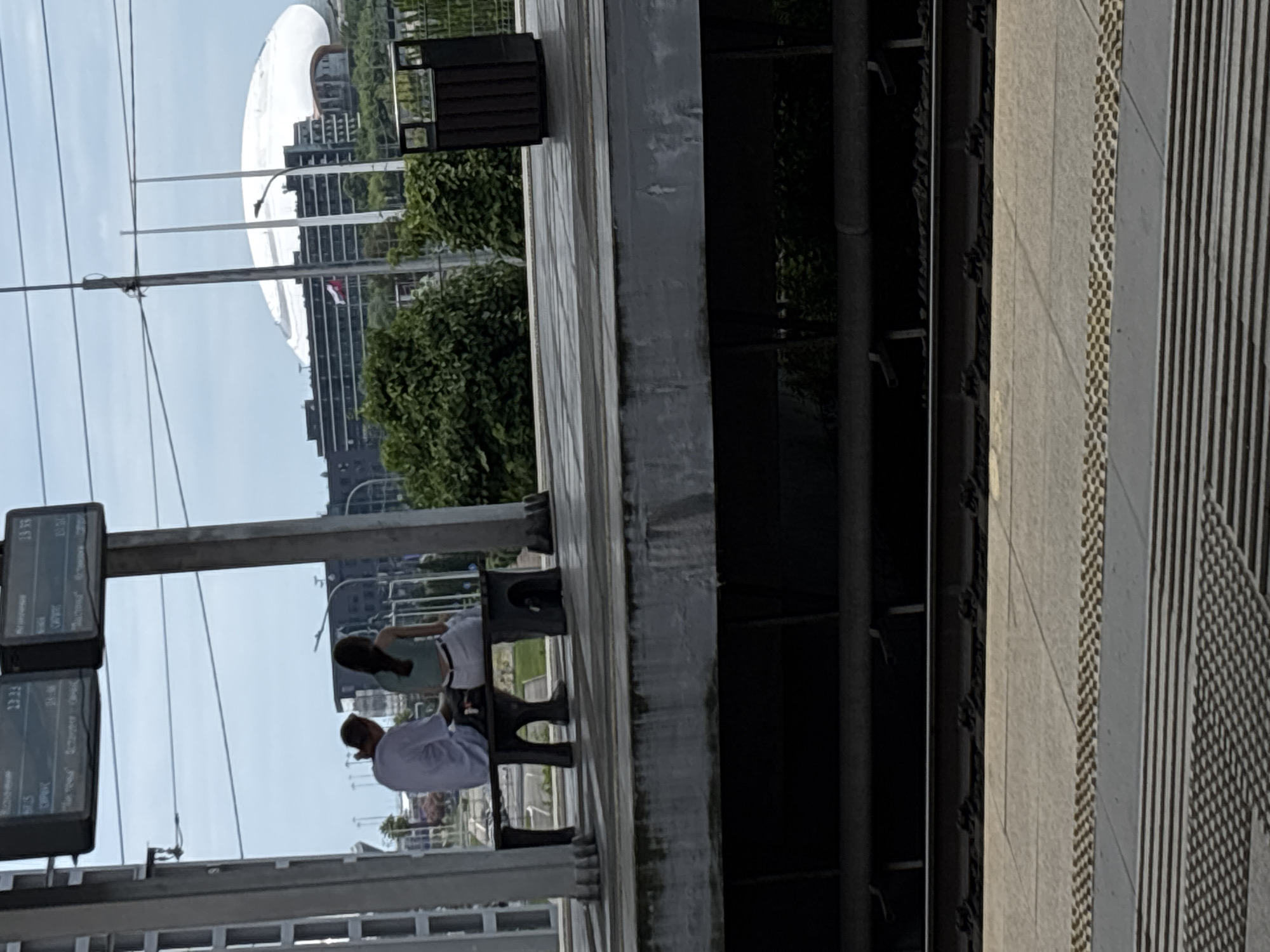 This is a medium shot, outdoor, daylight photograph taken at the Sirius train station in Sochi, Russia. Two people are seated on a dark-colored bench. The person on the left appears to be a male wearing a white t-shirt and dark pants. The person on the right is a female wearing a light green top and white pants. They are sitting close together and facing away from the camera, looking towards the train tracks. Behind them, a large, modern white building with a domed roof is visible in the distance, along with some greenery and other structures. The train station platform is visible in the foreground, with train tracks and tactile paving. The overall atmosphere is calm and ordinary.