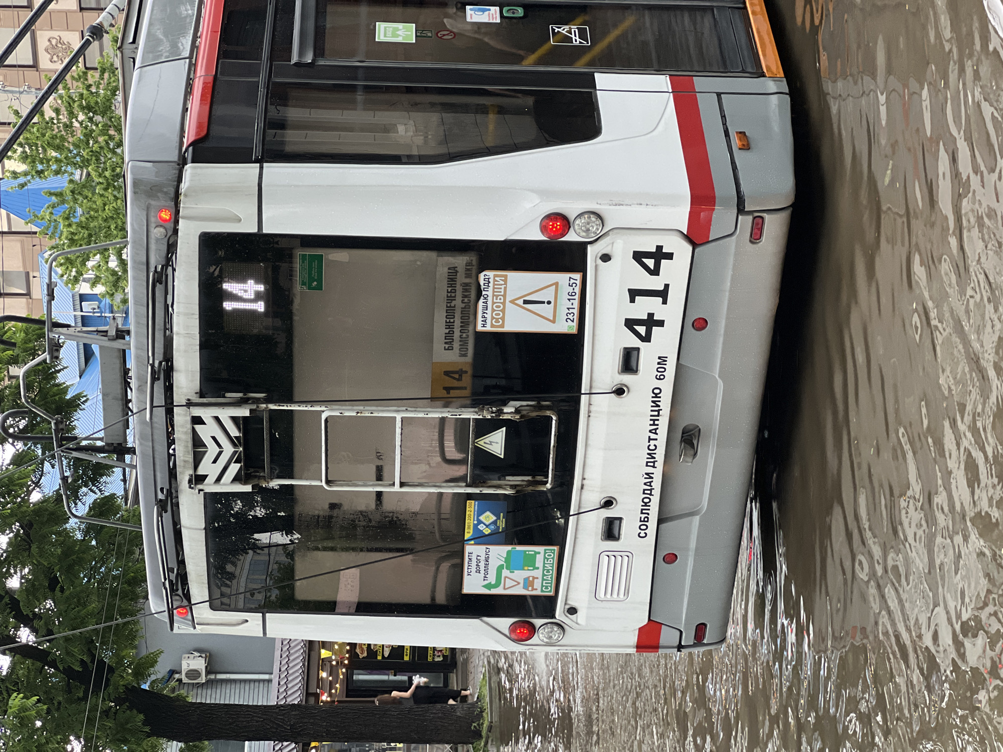 The image shows the rear of a white trolleybus with red and gray accents, numbered "14" on its destination display and "414" on its rear. The trolleybus is partially submerged in floodwater, indicating a flooded street. Several stickers are visible on the rear of the trolleybus, including a sign with an exclamation mark and text that appears to be in Russian, suggesting a traffic violation report. Another sticker requests drivers to yield to the trolleybus. Trees and buildings are visible in the background, with one building featuring a blue roof. The sky appears overcast. The scene is likely taking place in Krasnodar, Russia, given the Russian text on the trolleybus and the overall urban environment. There is a person walking on the sidewalk to the left of the trolleybus.