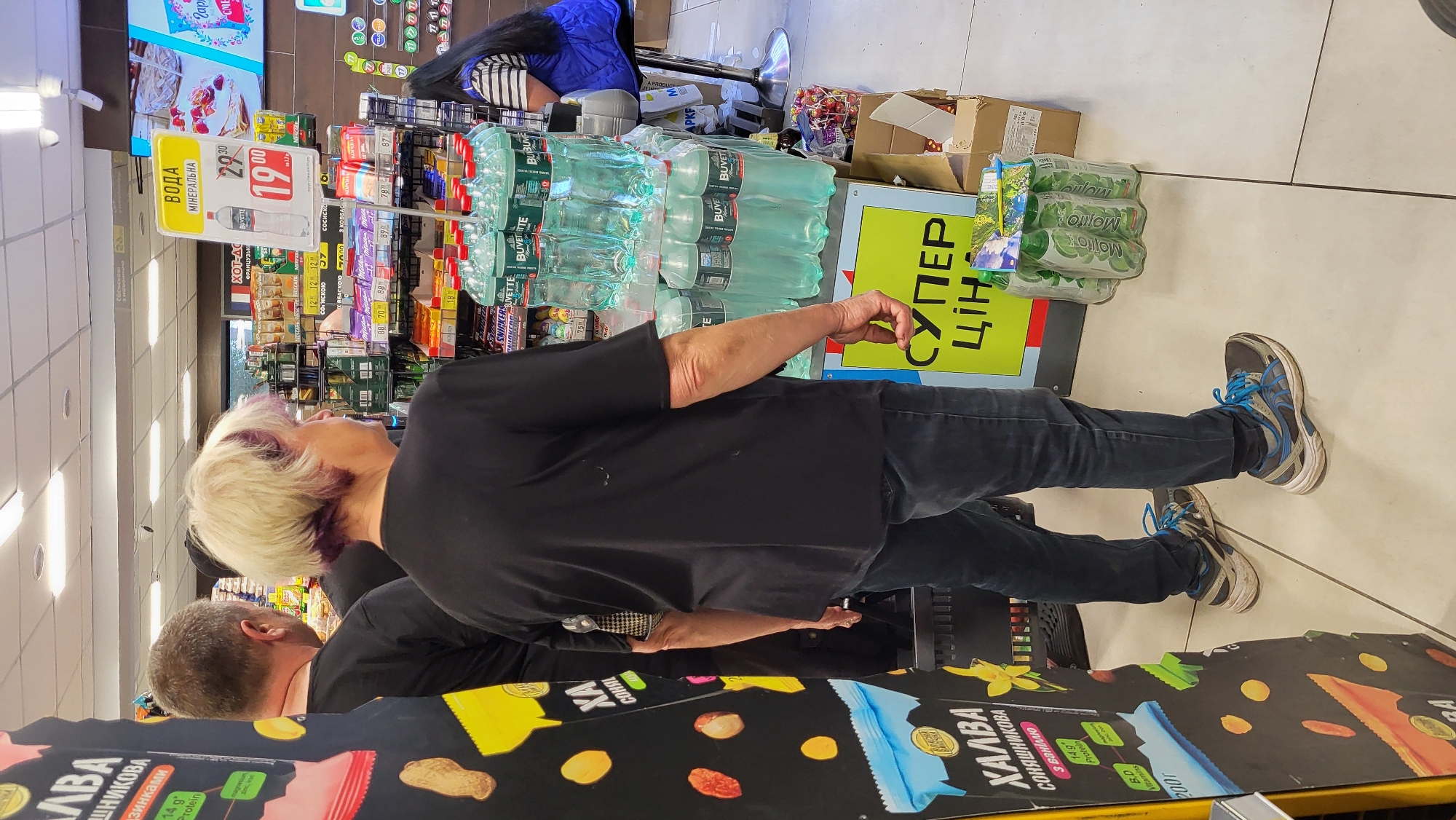 The image shows the interior of a supermarket, likely in the beverage aisle. In the foreground, a woman with blonde hair and purple streaks is standing with her back to the camera, facing a display of bottled water. She is wearing a black t-shirt and dark jeans, and her hair is styled in a short, choppy cut. To her left, a man wearing a black t-shirt is partially visible, also facing the beverage display.
The beverage display features multiple bottles of Buvette mineral water, neatly stacked on shelves. Above the bottles, a yellow sign with red lettering reads "ВОДА МІНЕРАЛЬНА" (Mineral Water) and shows a price of "29.90" with a sale price of "19.00". Another sign below the water bottles reads "СУПЕР ЦІНА" (Super Price) in large green lettering.
To the far left of the image, a portion of a black display stand is visible, featuring packaging with text in Cyrillic script, including the word "ХАЛВА" (Halva). Further back, other products and shelves are visible, along with a woman in a blue vest sitting on a stool. The floor is tiled. The overall lighting suggests an indoor retail environment.