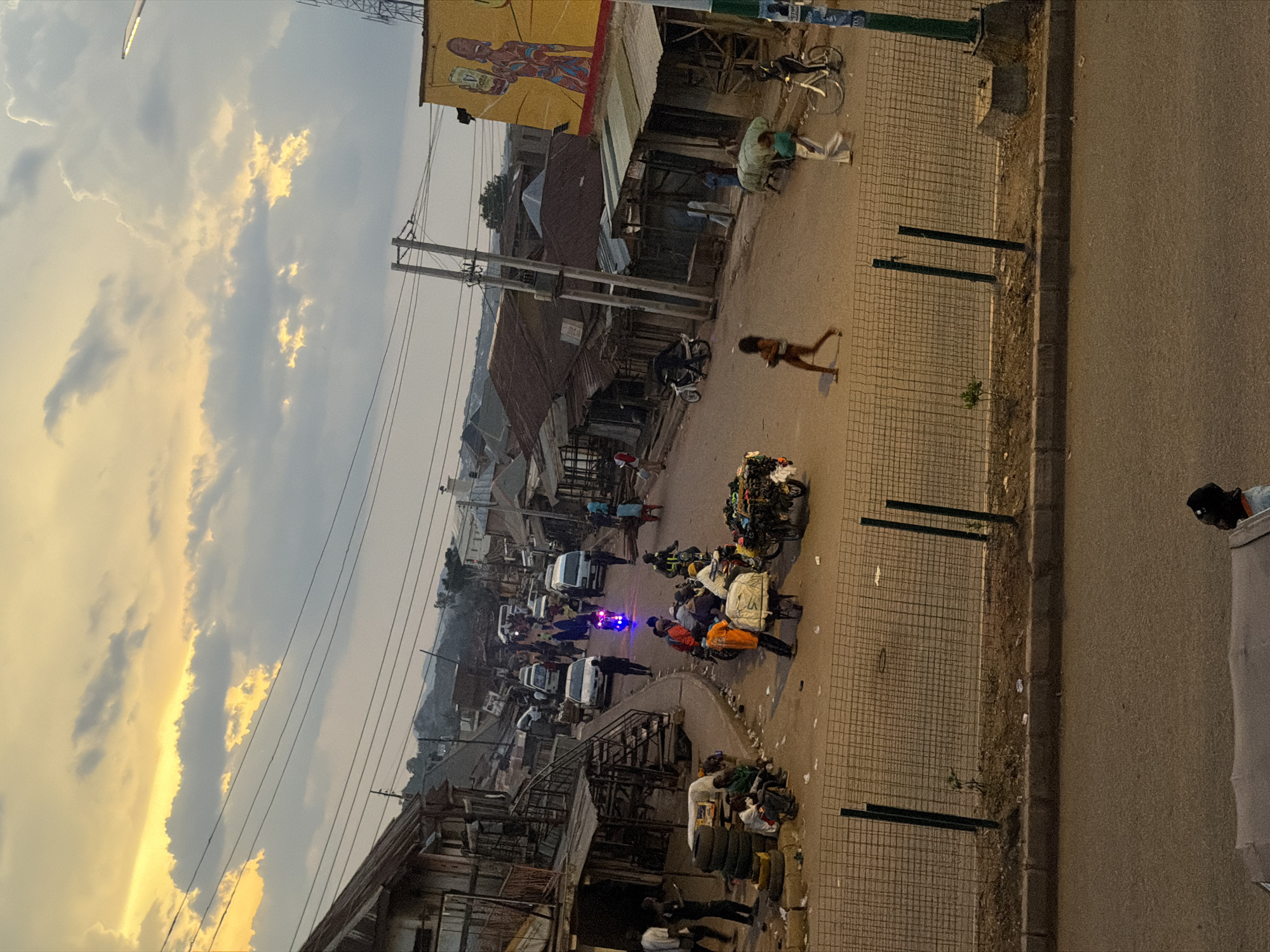 The image depicts a street scene in Bukuru, Nigeria, during what appears to be either dawn or dusk, with a dramatic sky filled with clouds illuminated by a golden hue. The street is lined with buildings, many of which have corrugated iron roofs, suggesting a bustling marketplace or residential area. A variety of vehicles, including cars and motorcycles, are present on the road, indicating daily activity. Several individuals are visible, engaged in various activities: some are walking, others are riding motorcycles, and a few appear to be involved in loading or unloading goods from carts. A prominent mural adorns the side of one building, adding a splash of color to the otherwise muted tones of the environment. The foreground features a wire fence, separating the road from a slightly elevated area with some vegetation and a concrete curb. The overall impression is one of a lively, working-class urban environment.