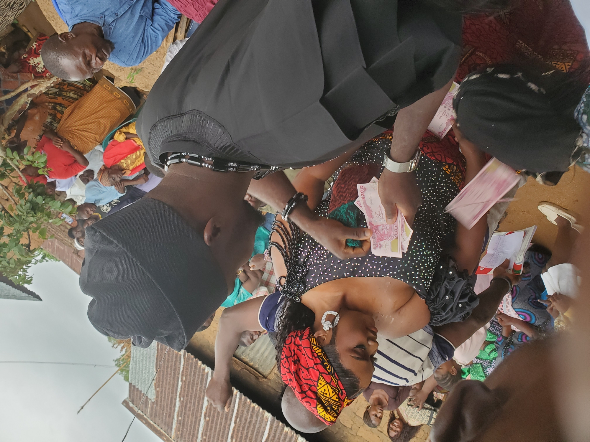 The image depicts an outdoor gathering, possibly in Jos, Nigeria, with several individuals in the foreground and background. In the center, a woman with a red and yellow headwrap is being handed money by a man wearing a black cap and a black robe with beaded embellishments. The woman is wearing a black dress adorned with white dots and has large silver earrings. Several other people are visible in the background, some seated and others standing. The environment appears to be a village or informal settlement, with corrugated iron roofs and earthy ground visible. The atmosphere suggests a social event or a transaction taking place.