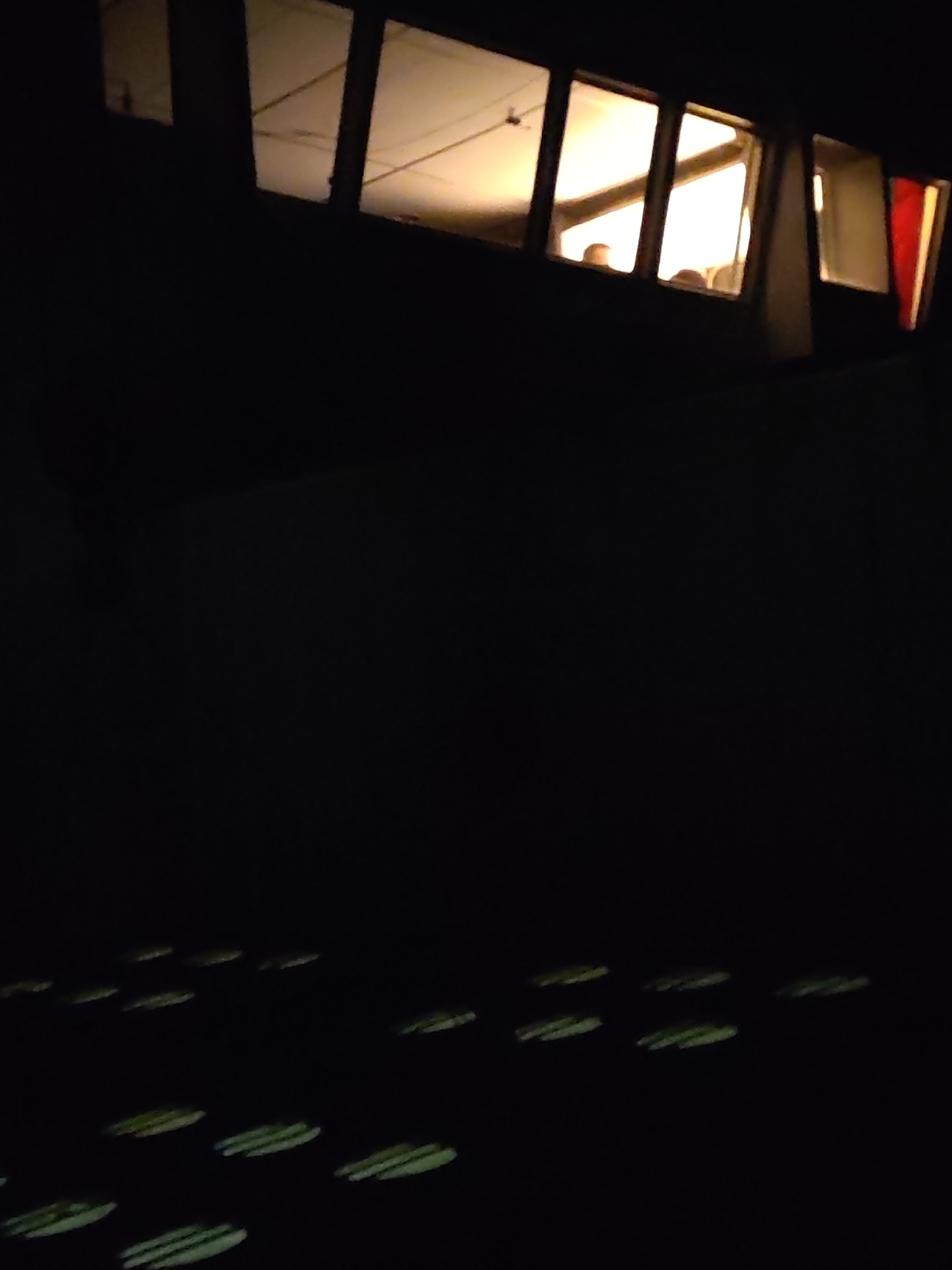 A nighttime image depicts a building's lit windows with people visible inside. The bottom part of the image is dark, with light patterns on the ground.