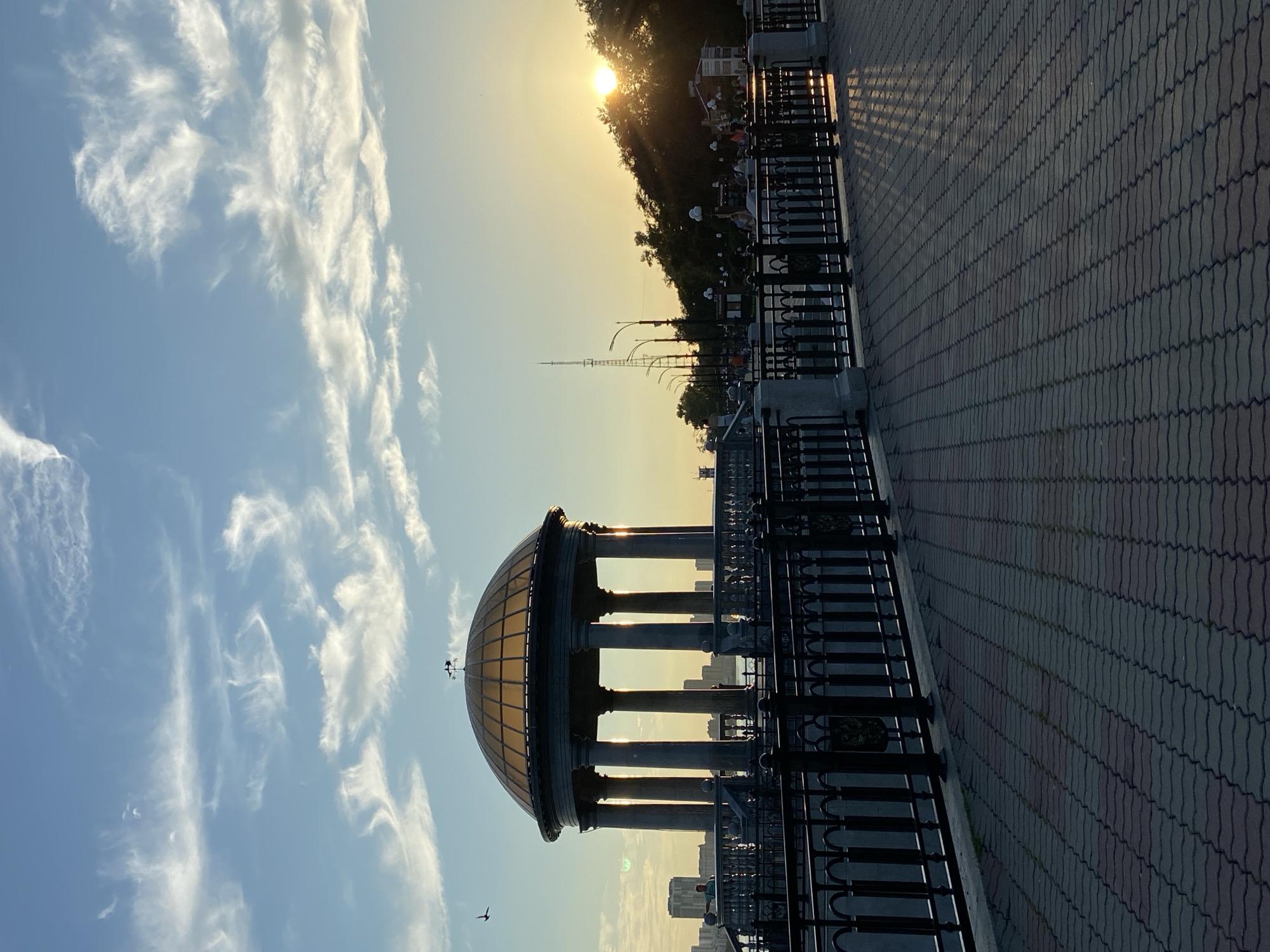 The image depicts a waterfront promenade in Blagoveshchensk, Russia, at sunset. A prominent rotunda with a golden dome, supported by classical columns, stands on the left side of the frame. A black metal railing runs along the edge of the promenade, separating it from the water. The ground is paved with interlocking bricks. In the background, a cityscape is visible across the river, and a dense line of trees forms a backdrop against the setting sun on the right. The sky is a blend of blue and orange hues, with wispy clouds illuminated by the setting sun. A few people are visible on the promenade, and a bird is flying in the sky. The overall scene conveys a peaceful and scenic atmosphere.