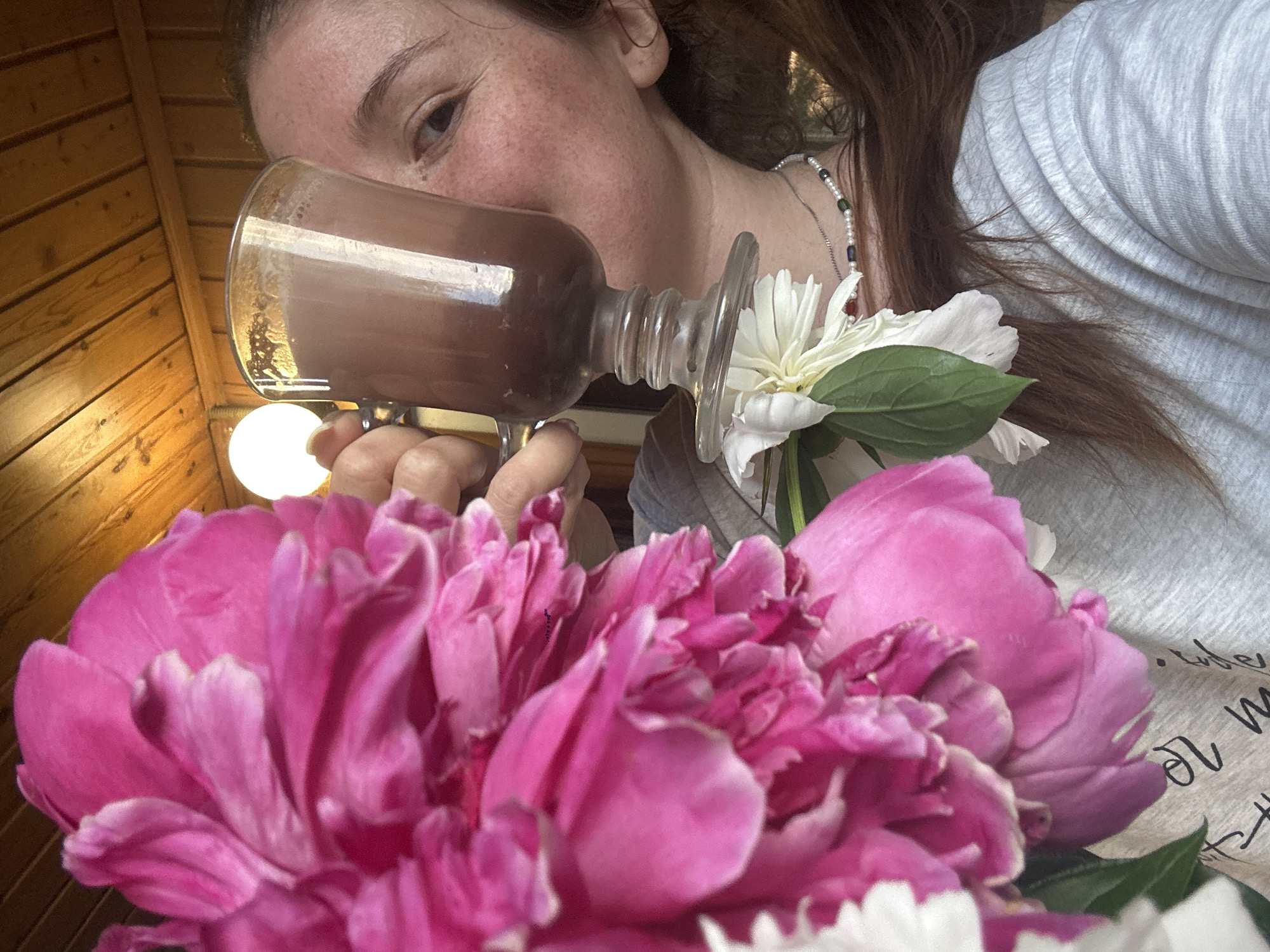 A close-up, medium shot captures a person holding a glass of dark liquid to their lips, partially obscured by a large pink peony flower in the foreground. The person, with light skin and freckles, has their right hand holding the glass and is looking towards the camera. They are wearing a light gray crew neck shirt with a black graphic design. A delicate pearl necklace with colorful beads is visible around their neck. Behind the person, a wooden-paneled ceiling with exposed beams is visible, along with a glowing light fixture. A small white flower with green leaves is positioned on the stem of the glass. The image is brightly lit, and the focus is on the person and the flowers, with the background slightly blurred. The scene appears to be indoors, possibly a cafe or a home, with a rustic aesthetic. The person's hair is reddish-brown and long, falling over their shoulder. The pink peony, with its abundant petals, dominates the left side of the frame, creating a soft, natural element. The overall impression is one of casual enjoyment and relaxation. The location is not specified, but it is implied to be a domestic setting.
