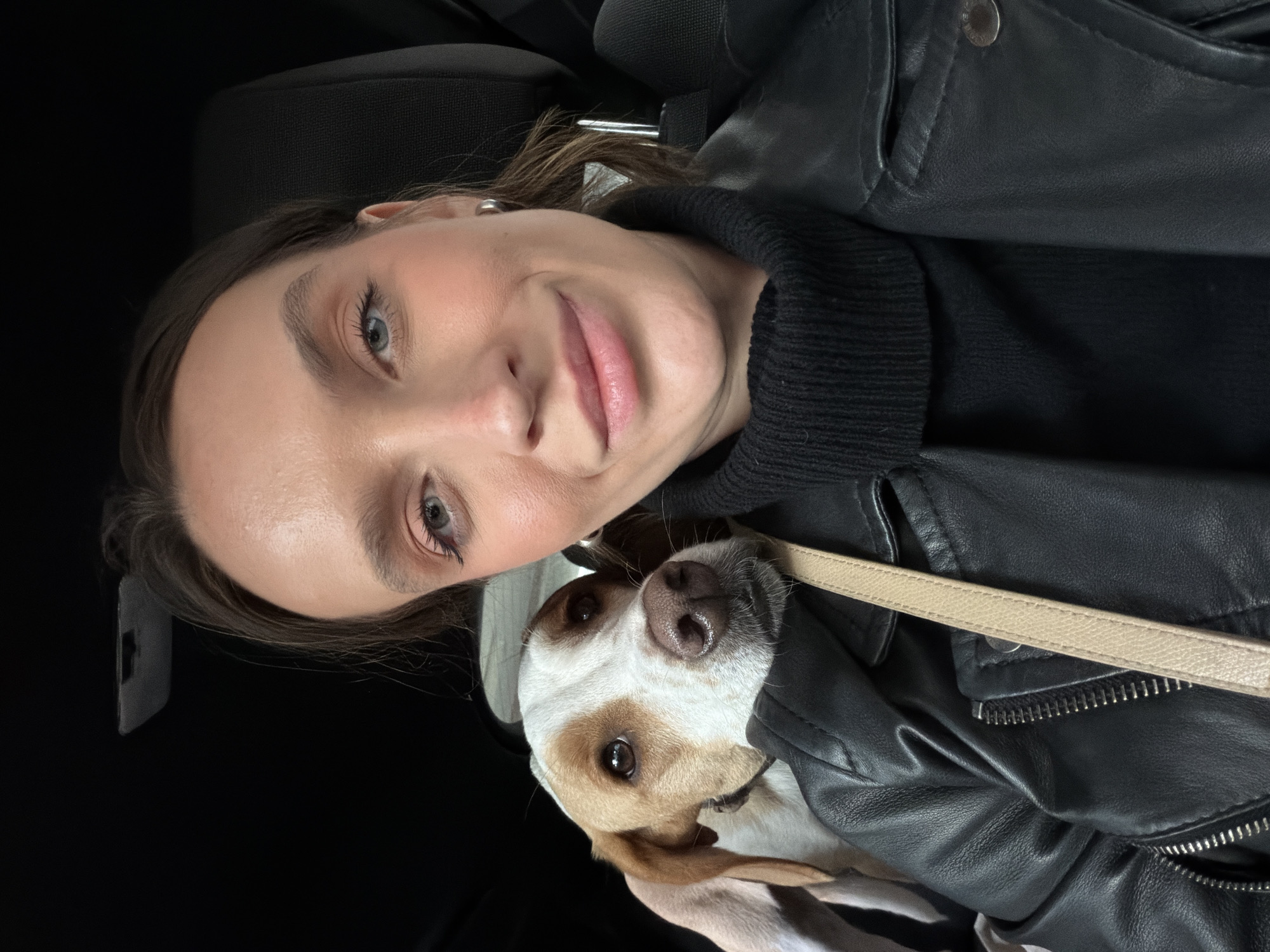The image displays an adult female and a medium-sized dog occupying the interior of a vehicle, in a scene situated in Budva, Montenegro. The female subject, positioned on the right, has fair skin, light brown hair, and blue eyes. She wears a black turtleneck sweater, a black leather jacket, and a silver hoop earring on her visible right ear. A light tan strap, likely from a bag, extends across her left shoulder. She exhibits a slight smile and maintains direct eye contact with the camera. The dog, positioned on the left side of the frame, features white and brown fur with brown eyes and a brown nose. It rests its head on the female's left shoulder, also looking towards the camera. The background consists of dark, undefined elements indicative of a car interior, including a black headrest behind the female.
