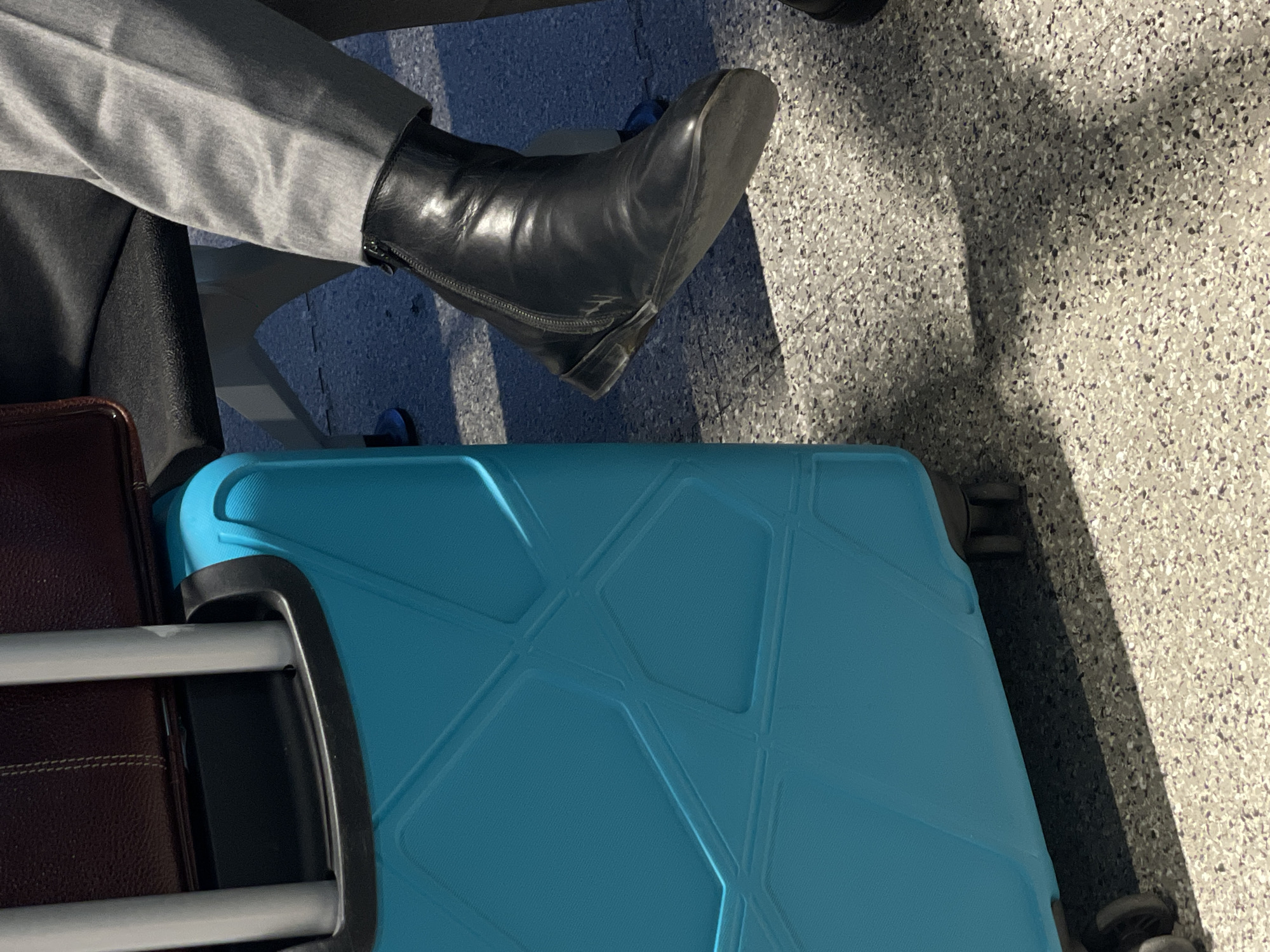 A top-down shot shows a seated person with a black boot, gray pants, and a black jacket. The person is seated next to a blue suitcase with a geometric pattern and a brown suitcase. The ground has a speckled, gray and white surface. The location is indeterminate; there is no verifiable evidence to place the scene in Tashkent, Uzbekistan.