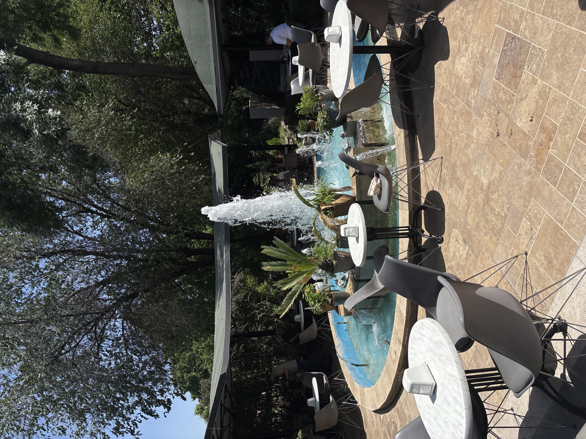 The image depicts an outdoor seating area of a cafe with a central water fountain in Yerevan, Armenia. The fountain is surrounded by turquoise water and has a spray of water in the air. Around the fountain, there are several tables with modern gray chairs and white marble tabletops. Potted plants, including a palm, are placed around the fountain.
In the background, there are shaded seating areas covered by awnings, where several patrons are seated at tables. One man in a white shirt is visible sitting at a table in one of the shaded areas. Trees and foliage surround the cafe, creating a natural and serene atmosphere. The ground is paved with light brown tiles. The overall impression is a pleasant outdoor dining setting.